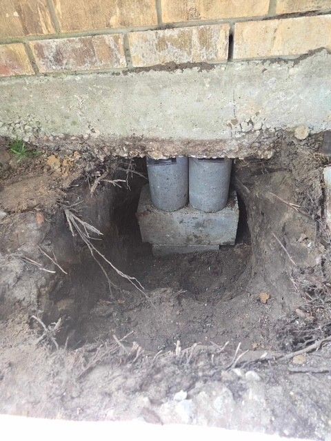 Hole dug in dirt reveals concrete base supporting two gray concrete posts beneath a brick structure