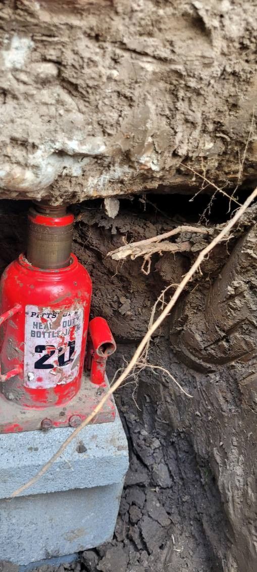 A red 20-ton hydraulic jack supporting a dirt-covered structure in an excavation