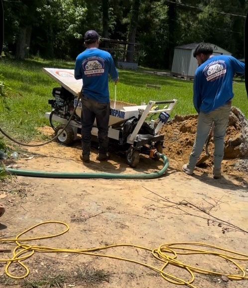 Two men in blue shirts operate machinery outdoors near a dirt pile and a hose