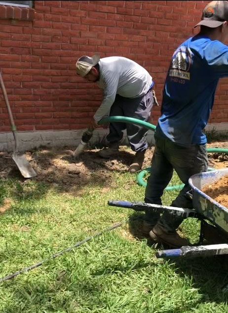 Two construction workers laying a green hose near a brick wall and grass