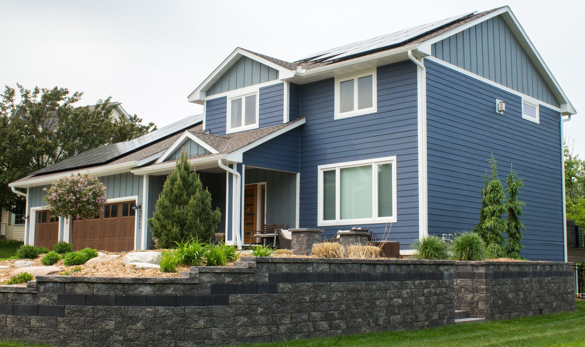 Two-story house with blue siding, solar panels, and a tiered retaining wall in a yard.