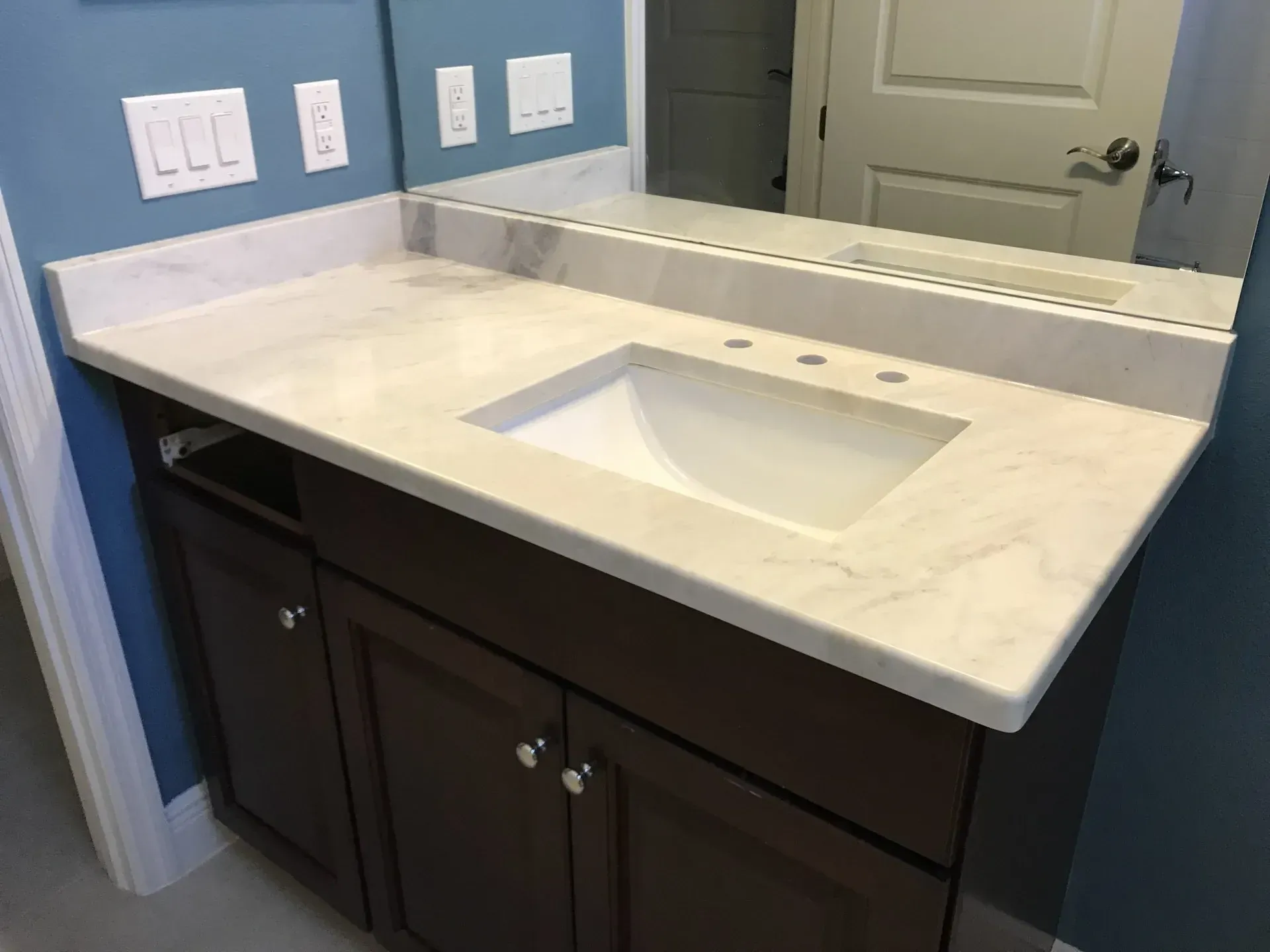 Modern bathroom vanity with white countertop, rectangular sink, dark cabinets, and large mirror.