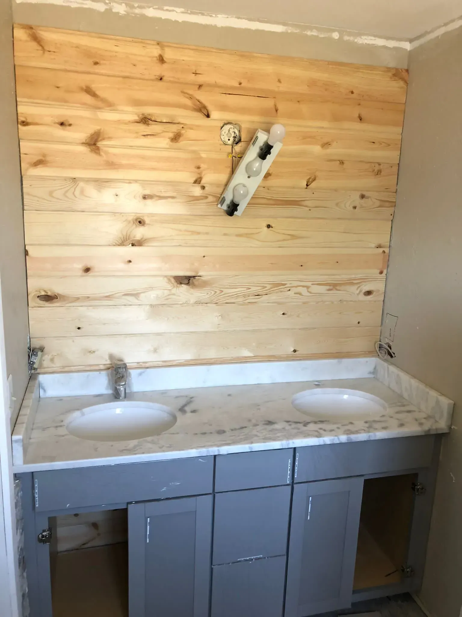 Partially finished bathroom vanity with double sinks, gray cabinets, and wood-paneled wall behind it