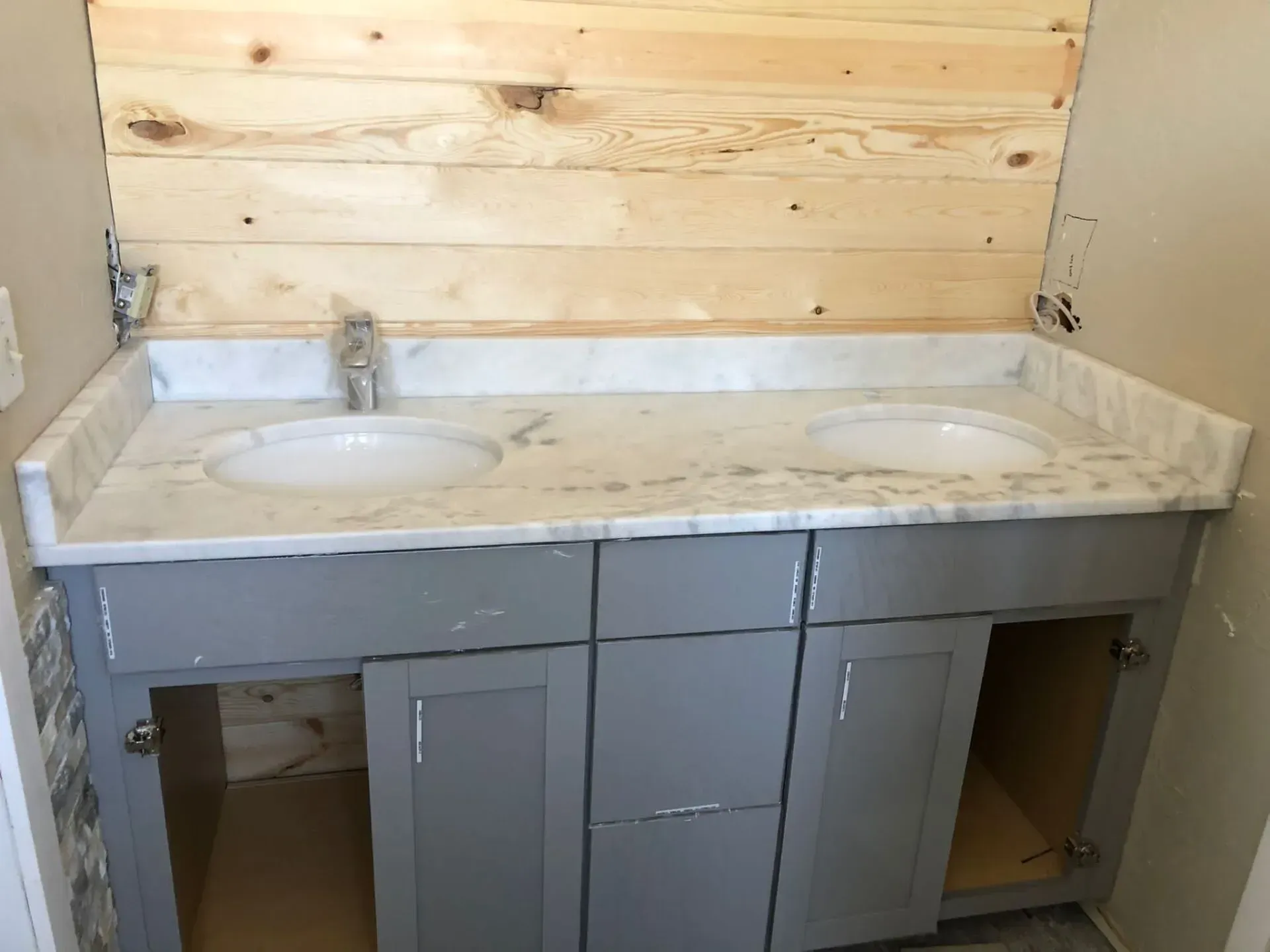 Unfinished bathroom vanity with marble countertop, double sinks, and gray cabinet doors missing beneath them