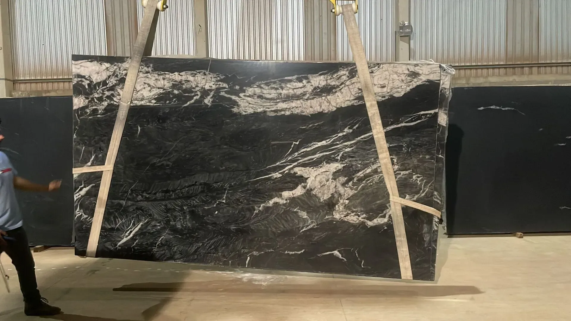 Large black-and-white stone slab suspended by straps in an industrial warehouse.