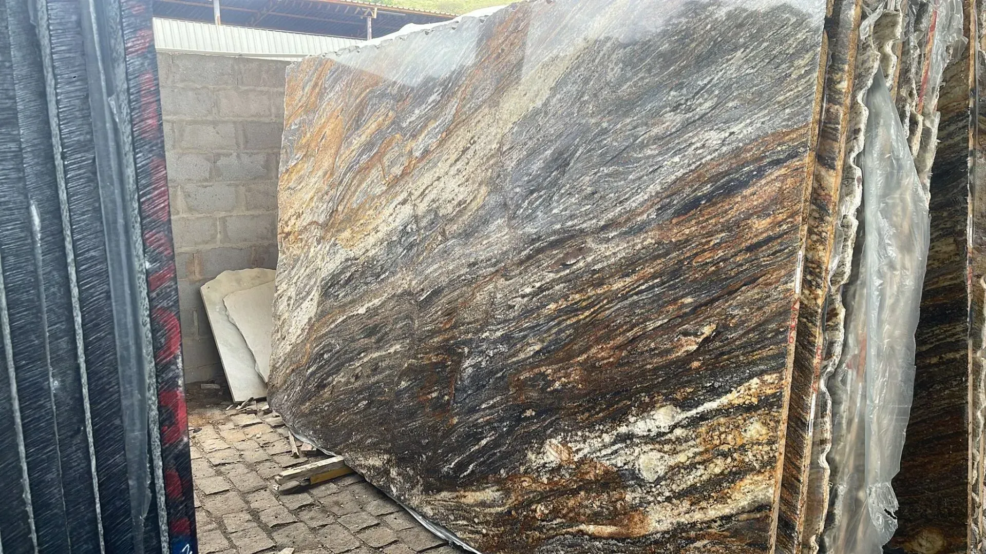 Large polished stone slabs stacked in an industrial yard, showing gray, brown, and cream veining.