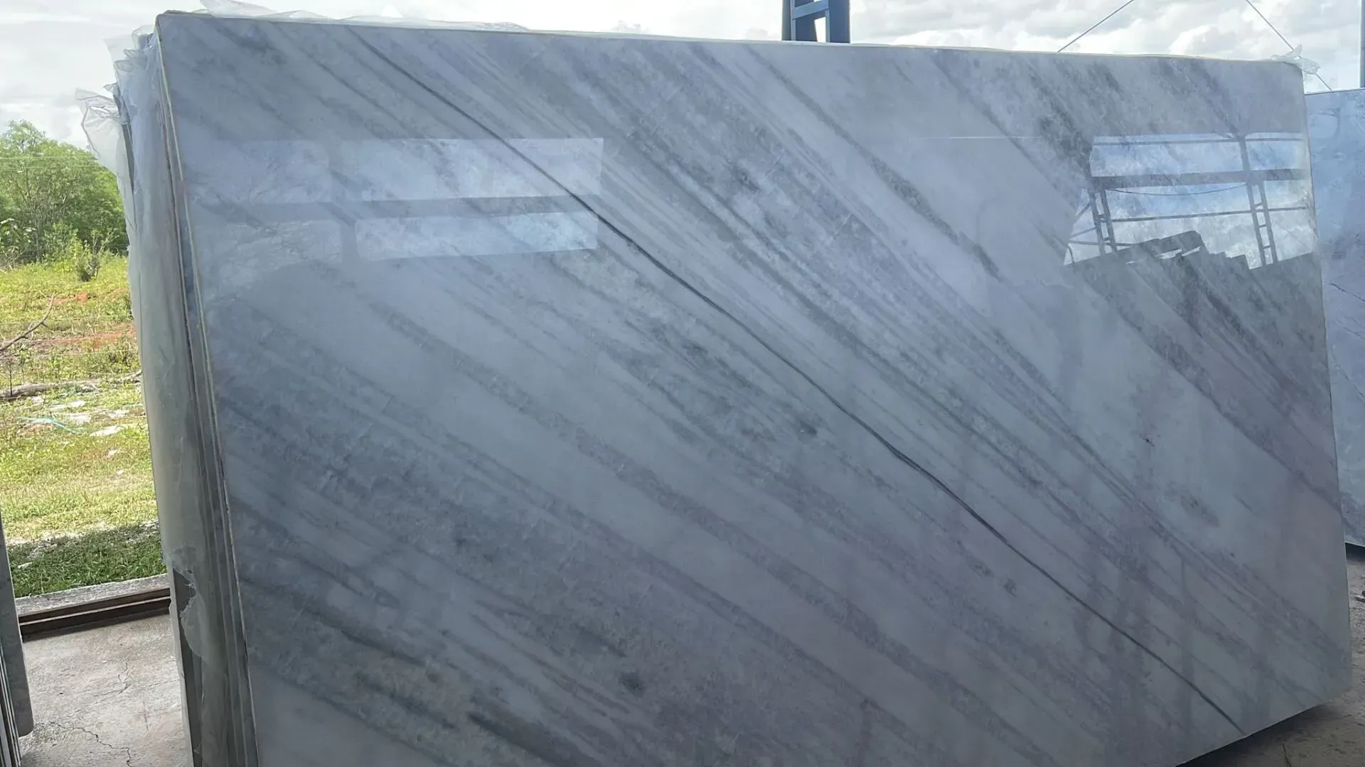 Large gray marble slab wrapped in plastic, leaning outdoors near grass and a metal frame.