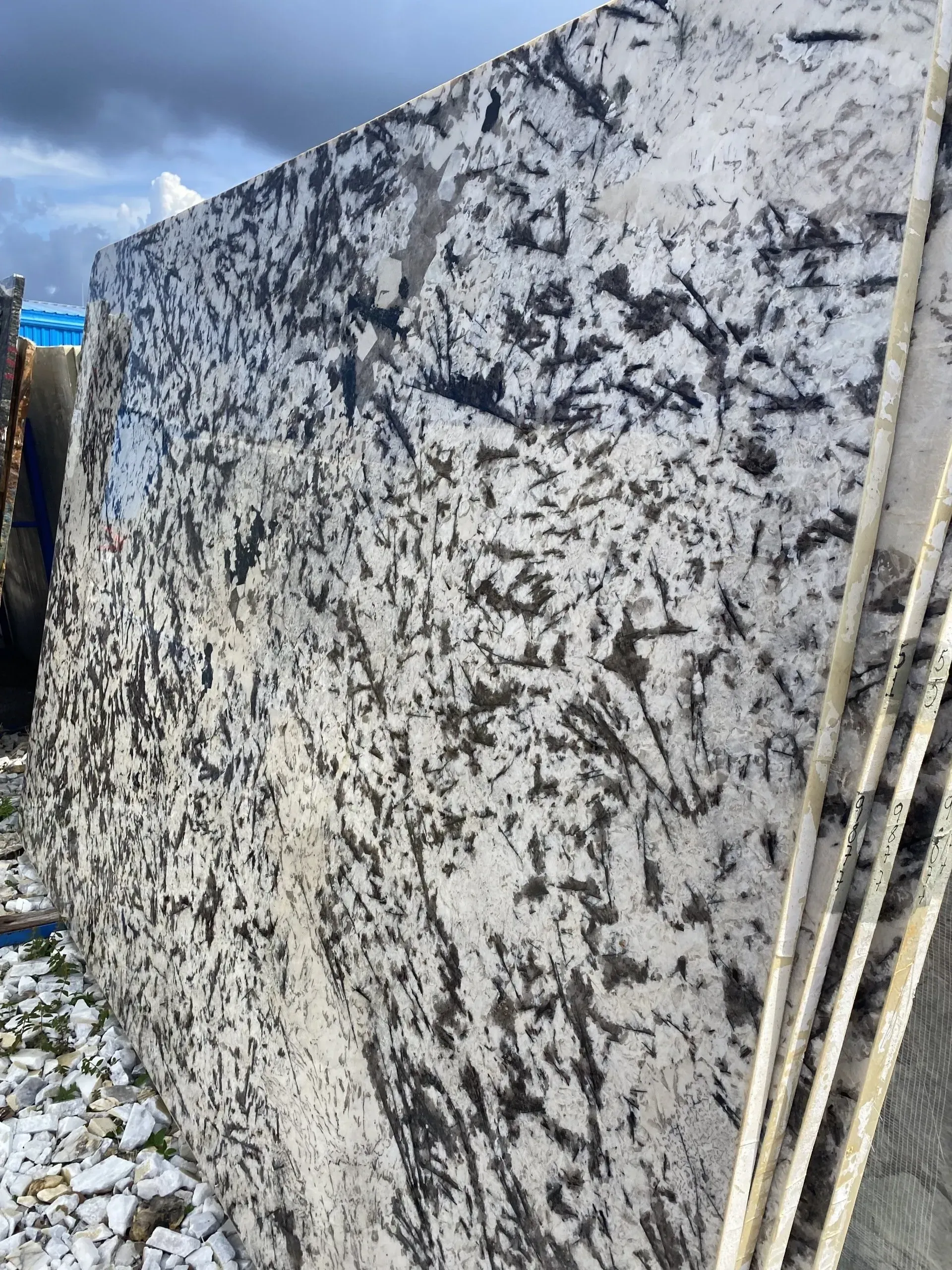 Large speckled granite slab outdoors with blue sky and stacked poles beside it