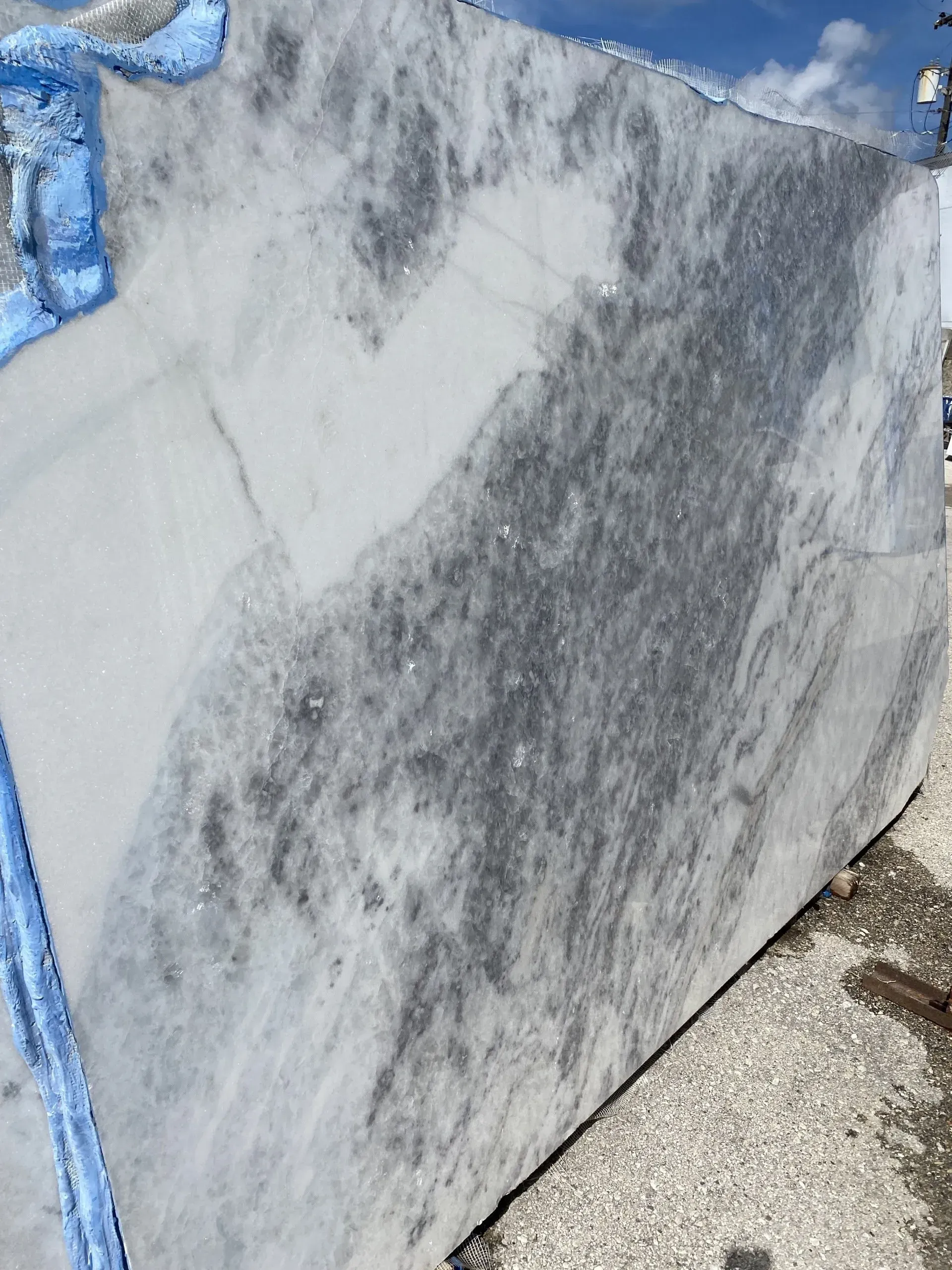 Large marble slab with gray veining leaning outdoors on gravel under a blue sky.