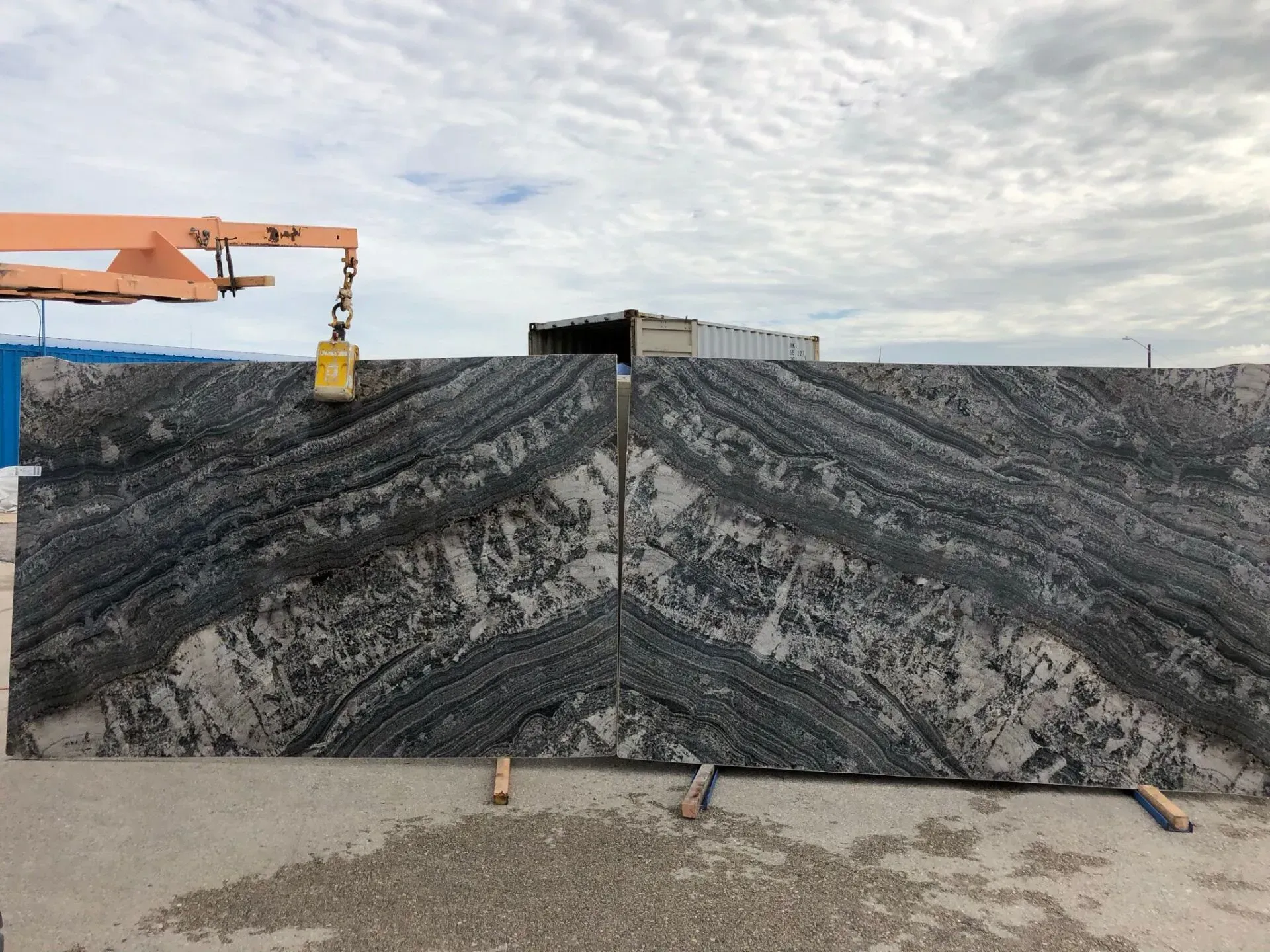 Large black stone slabs at a construction site, with a crane lifting one slab under a cloudy sky