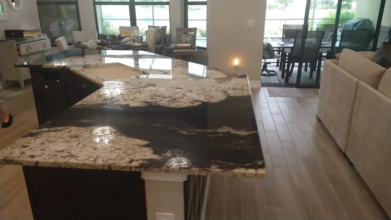 Modern kitchen with black granite island, barstools, beige sofa, and large windows overlooking a patio