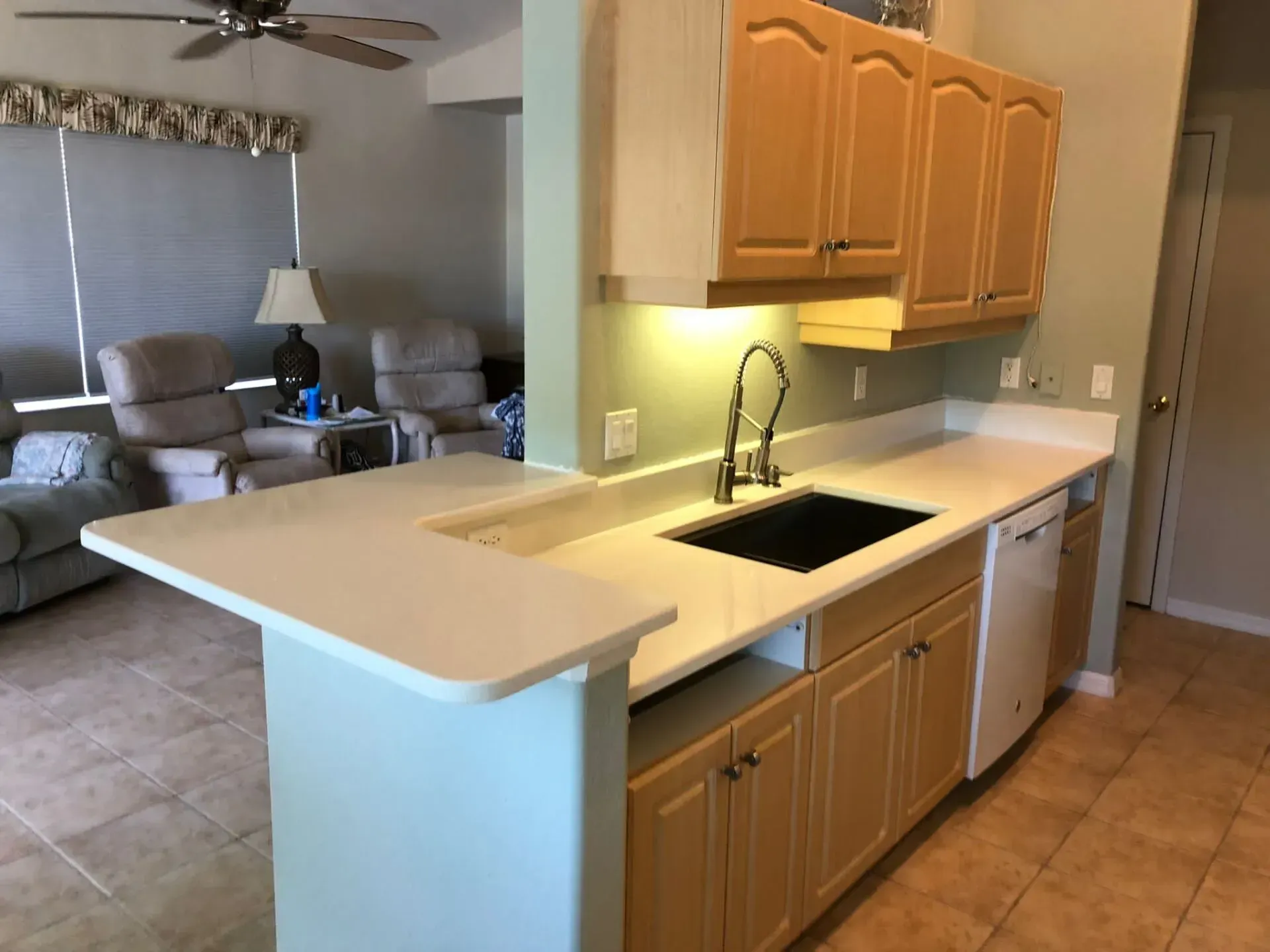 Kitchen with white island countertop, black sink, and wood cabinets opening to a living room.