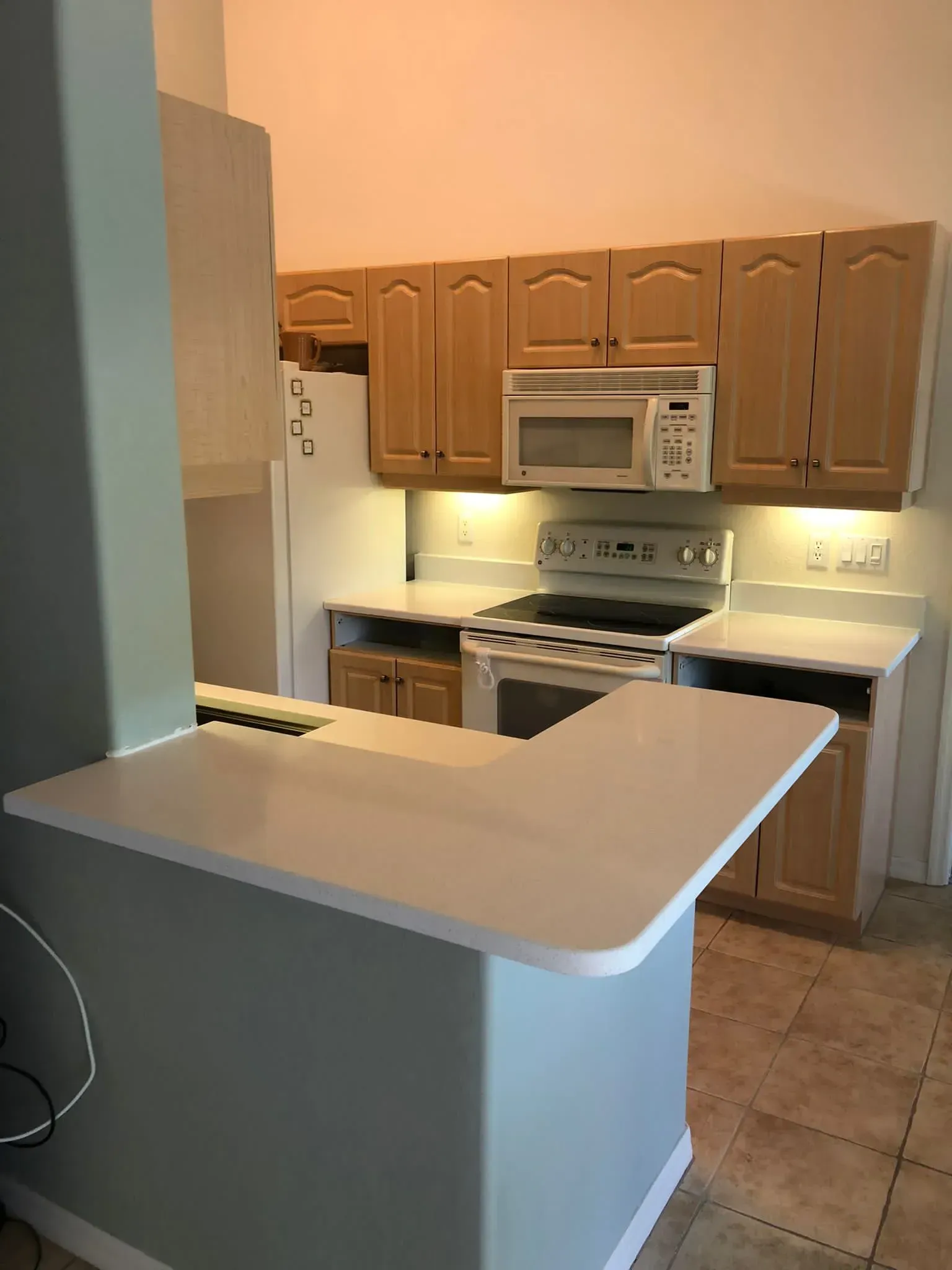 Small kitchen with tan cabinets, white appliances, and a light-colored breakfast bar in the foreground