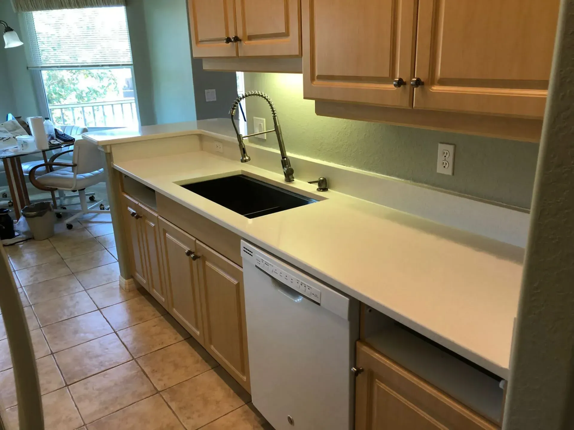 Kitchen with tan cabinets, white countertop, black sink, and a dishwasher by a window.