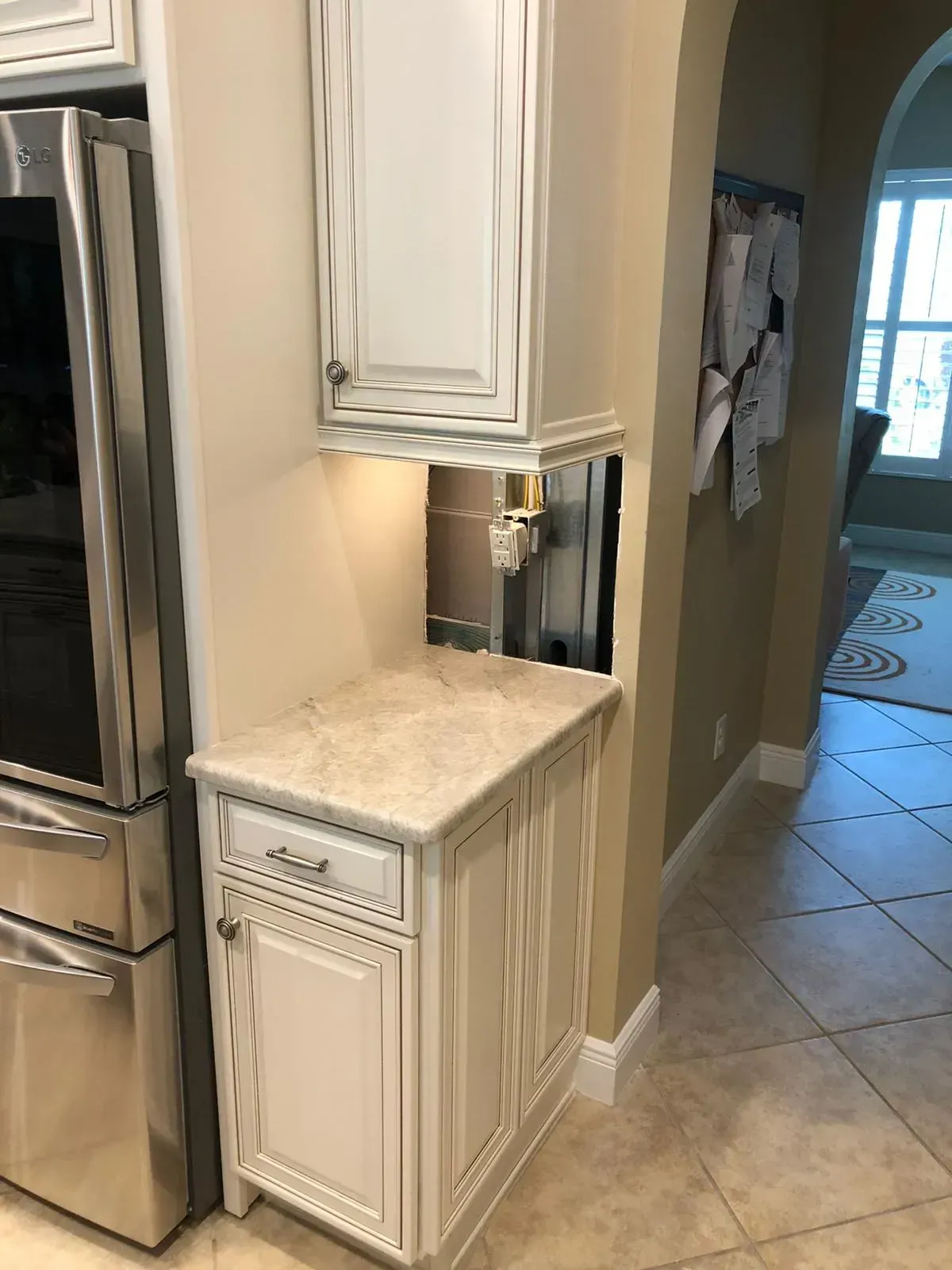White kitchen cabinet with granite countertop beside stainless steel fridge and tiled hallway.