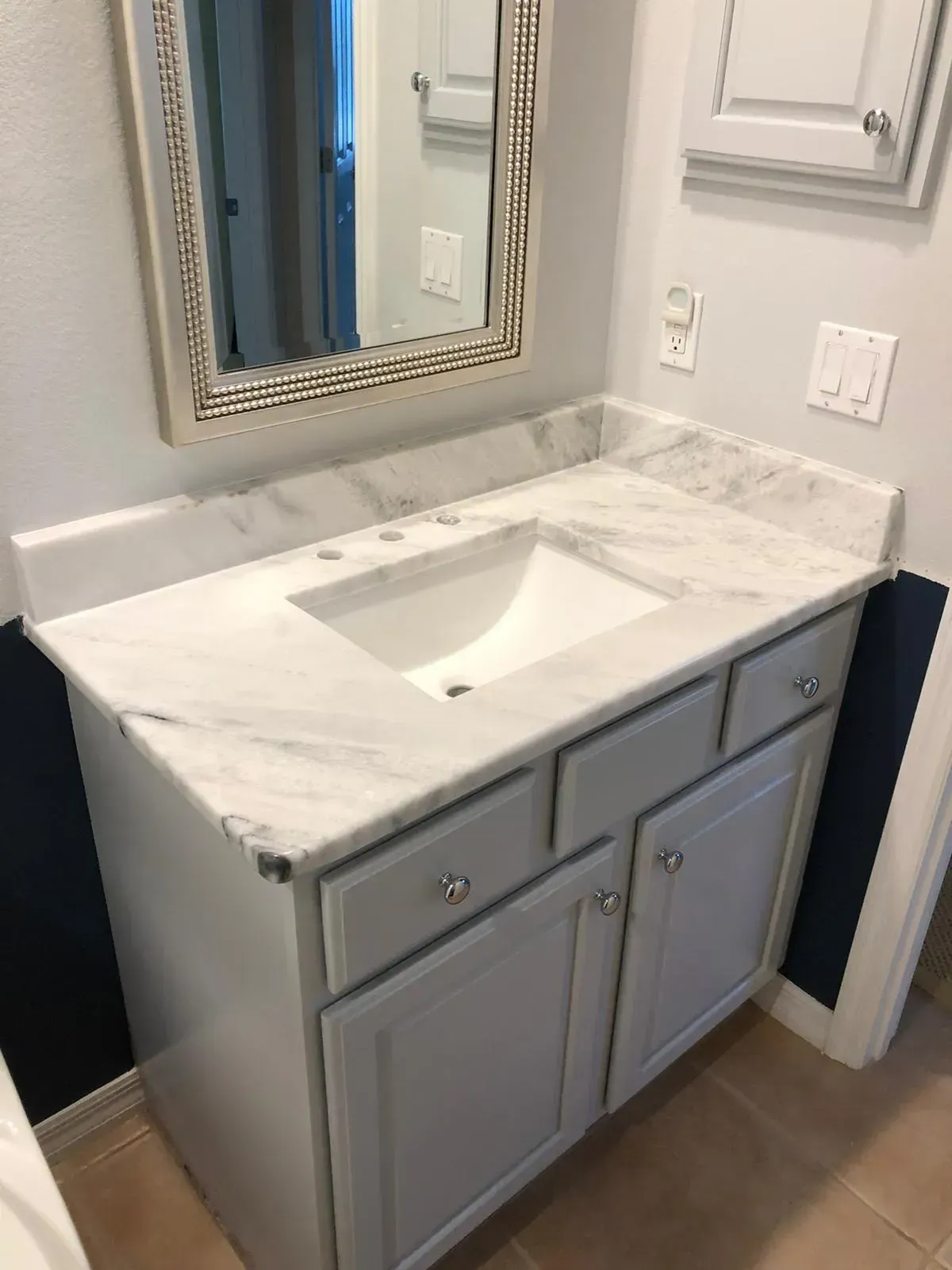 White bathroom vanity with marble countertop, sink, mirror, and gray cabinets against light walls.