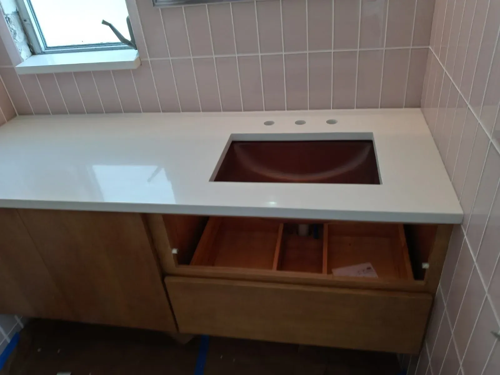 Modern bathroom vanity with white countertop, square sink cutout, and wood cabinets beneath a window.