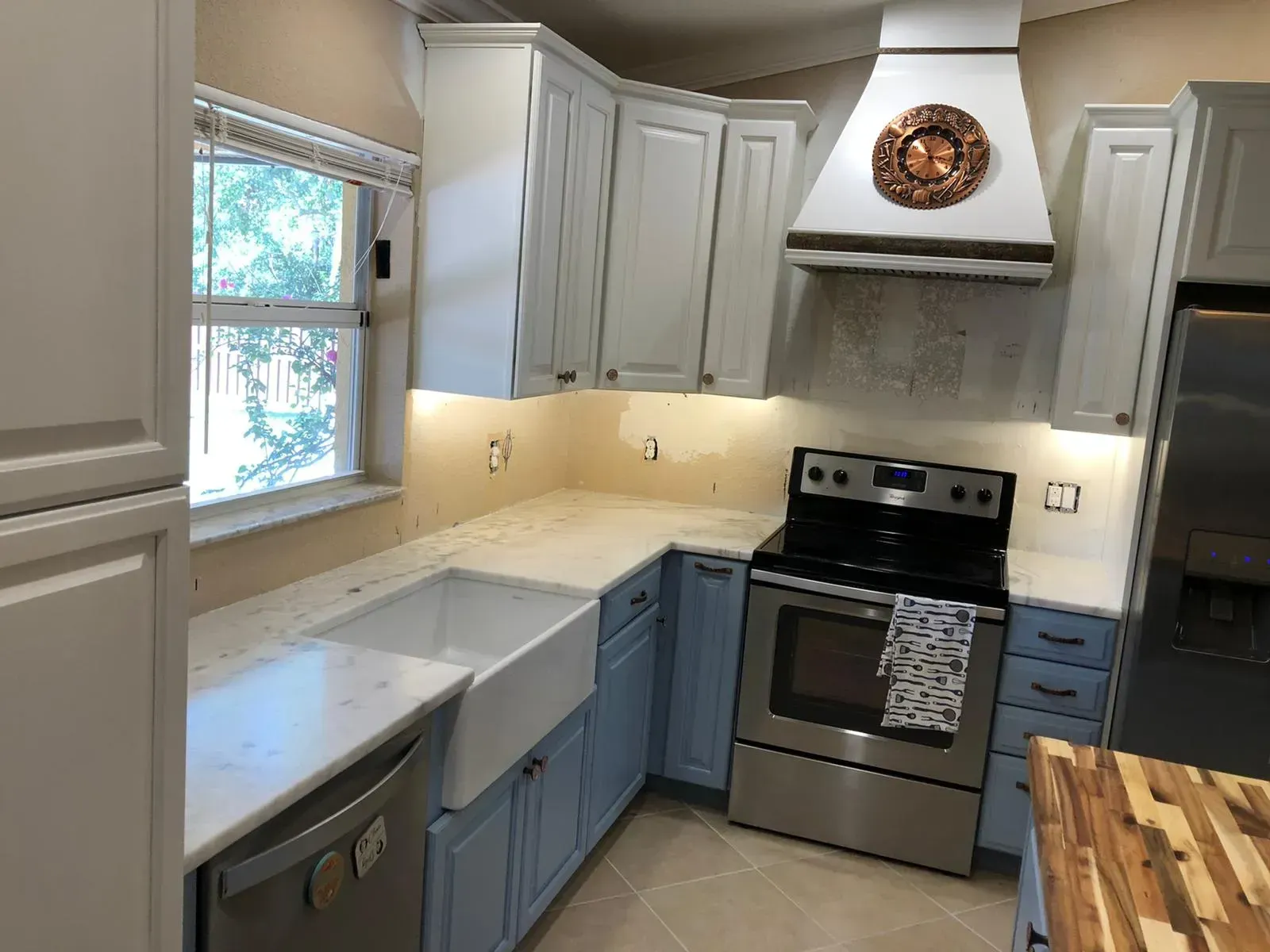 Bright kitchen with blue lower cabinets, white countertops, stainless stove, and window by the sink.