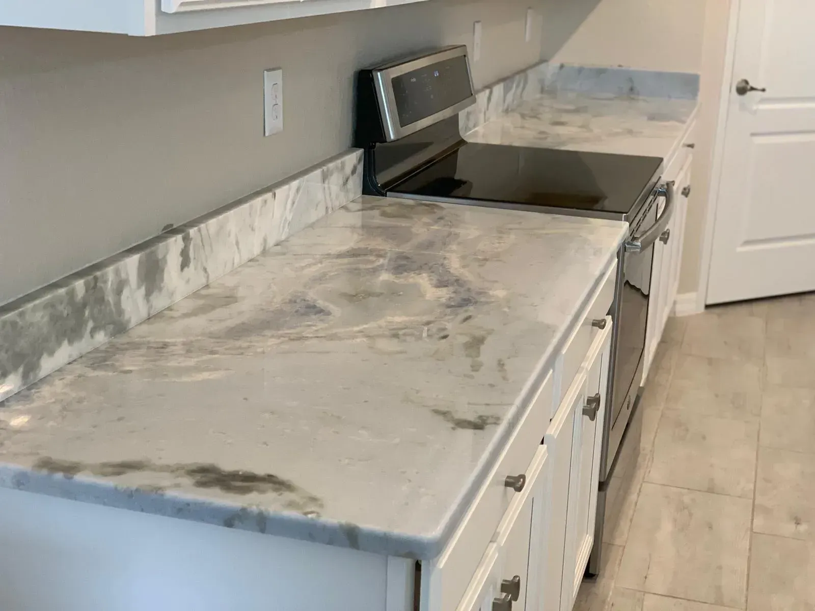 Kitchen countertop with cloudy white marble-like surface and matching backsplash next to a stove.
