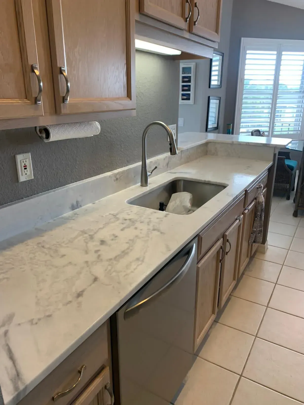 Kitchen with marble countertop, sink, stainless dishwasher, wood cabinets, and tiled floor beside a window.