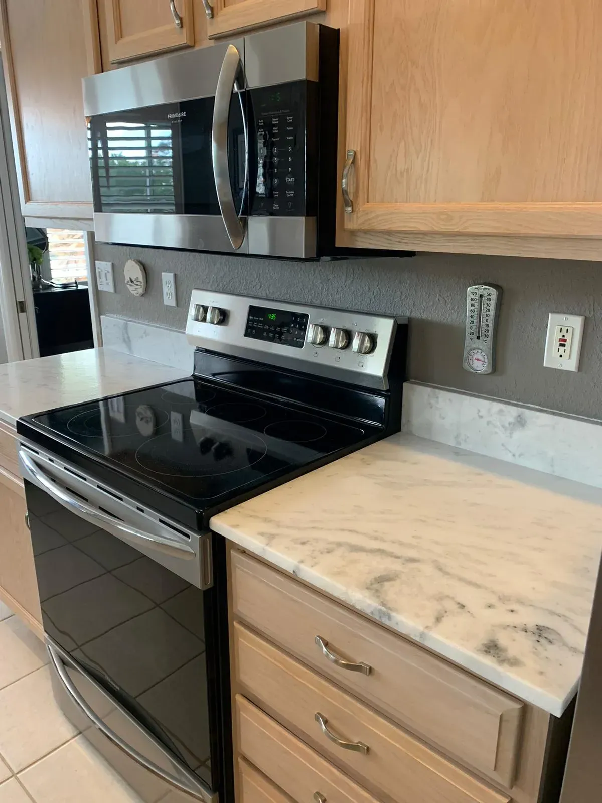 Kitchen with black stove, stainless microwave, and light wood cabinets beside a marble countertop.