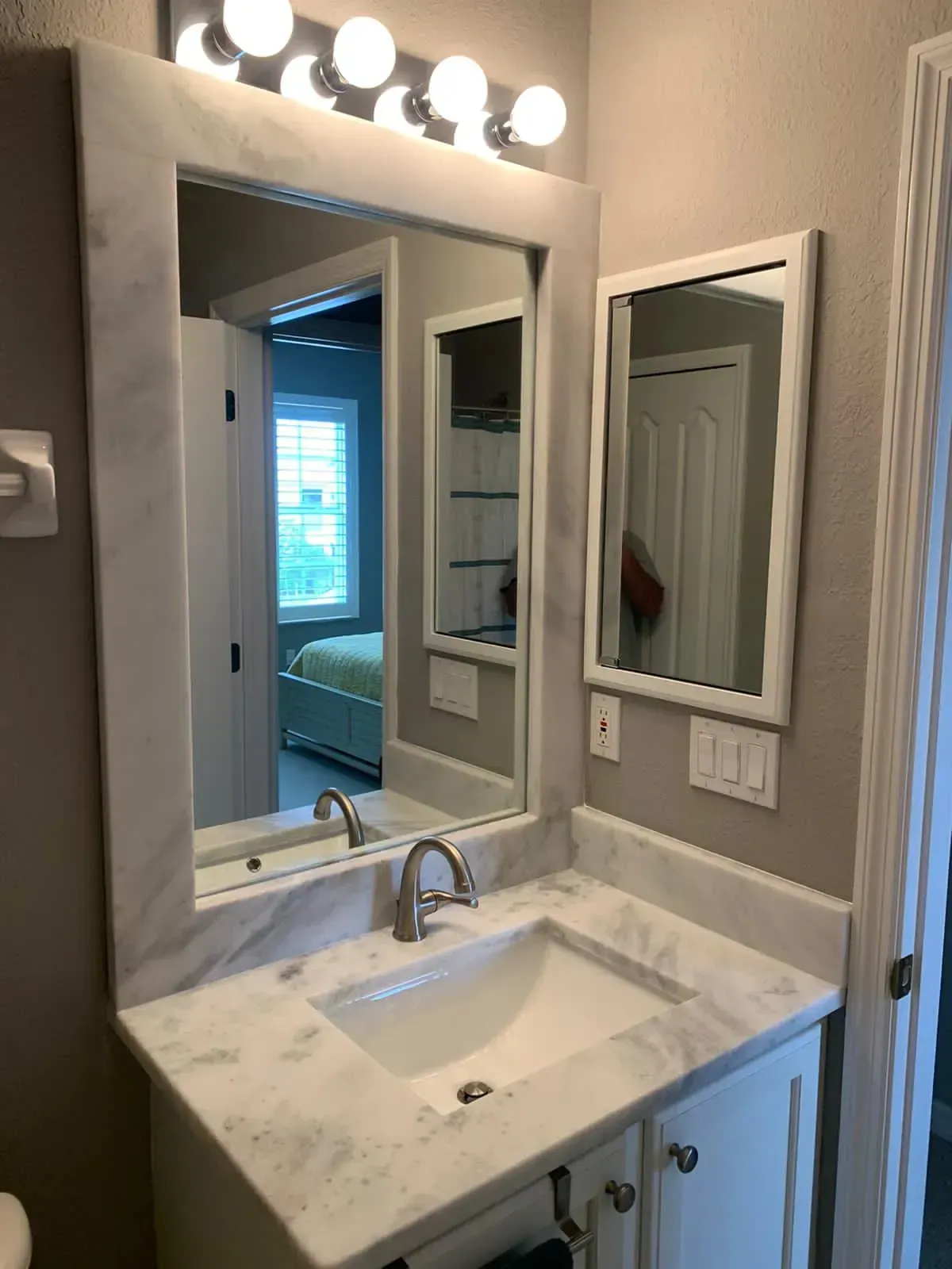 Small bathroom vanity with marble sink, mirror, wall lights, and an open doorway reflected in the mirror