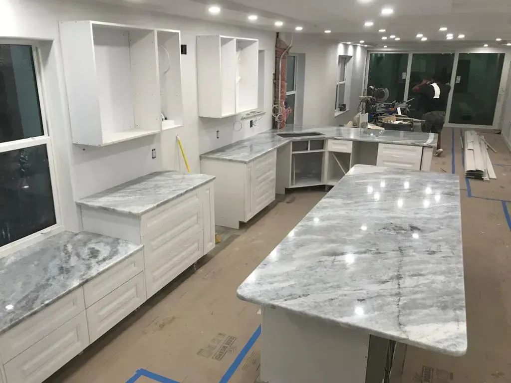 Unfinished kitchen remodel with white cabinets, gray marble countertops, and island under bright ceiling lights.
