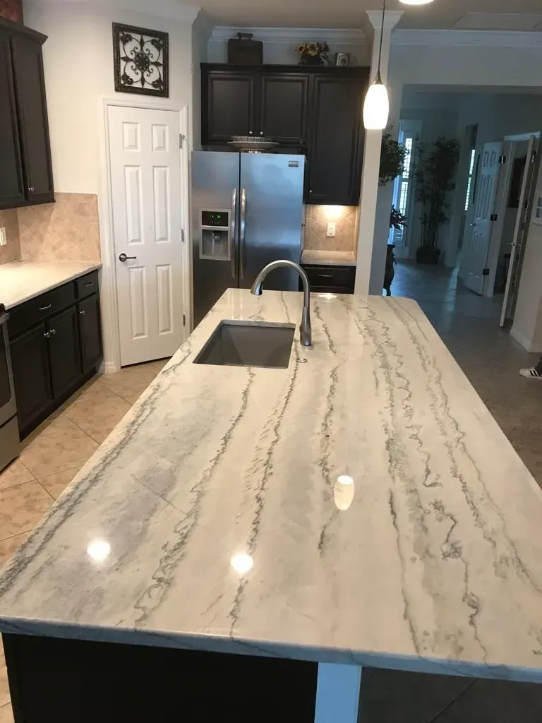 Modern kitchen with white marble island, stainless fridge, dark cabinets, and a sink under pendant lights