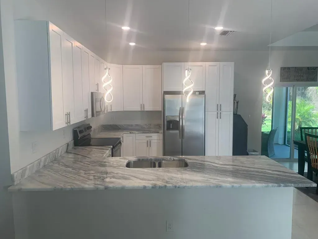Modern kitchen with white cabinets, marble island, stainless fridge, and large windows.