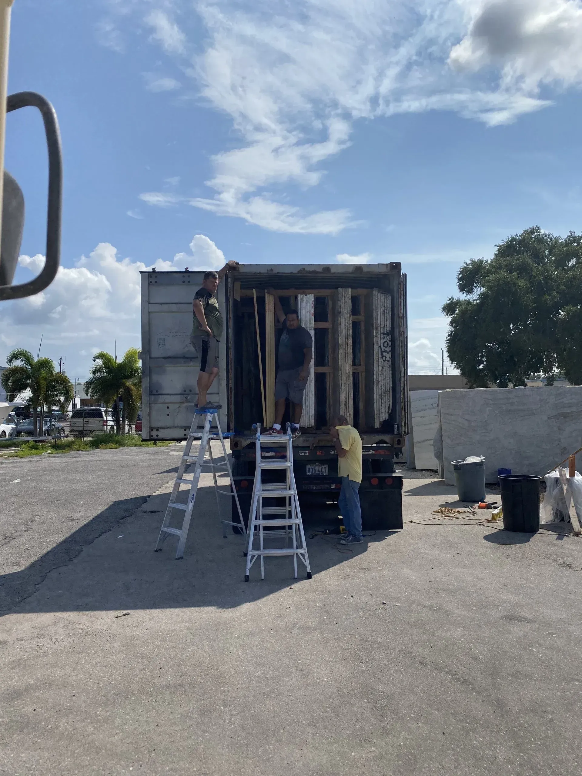 A small work trailer with ladders parked on a gravel lot under a bright, partly cloudy sky.