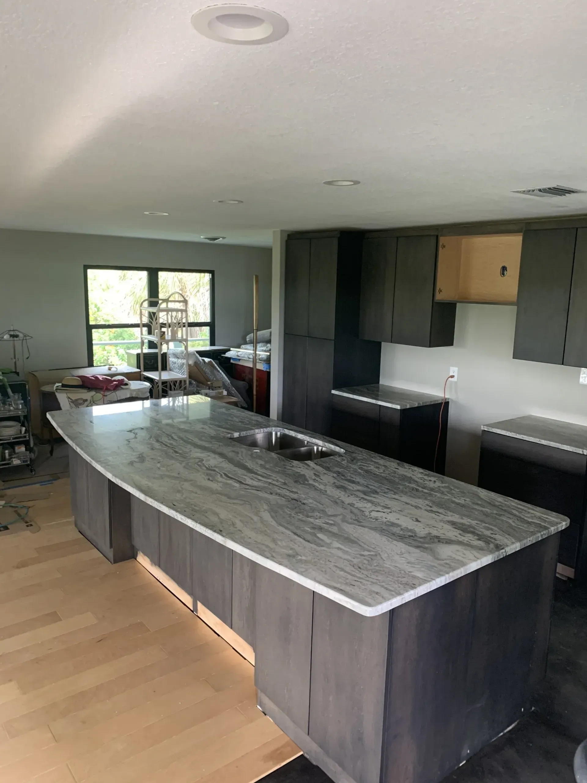 Modern kitchen with large marble island, dark cabinets, wood floor, and window view.