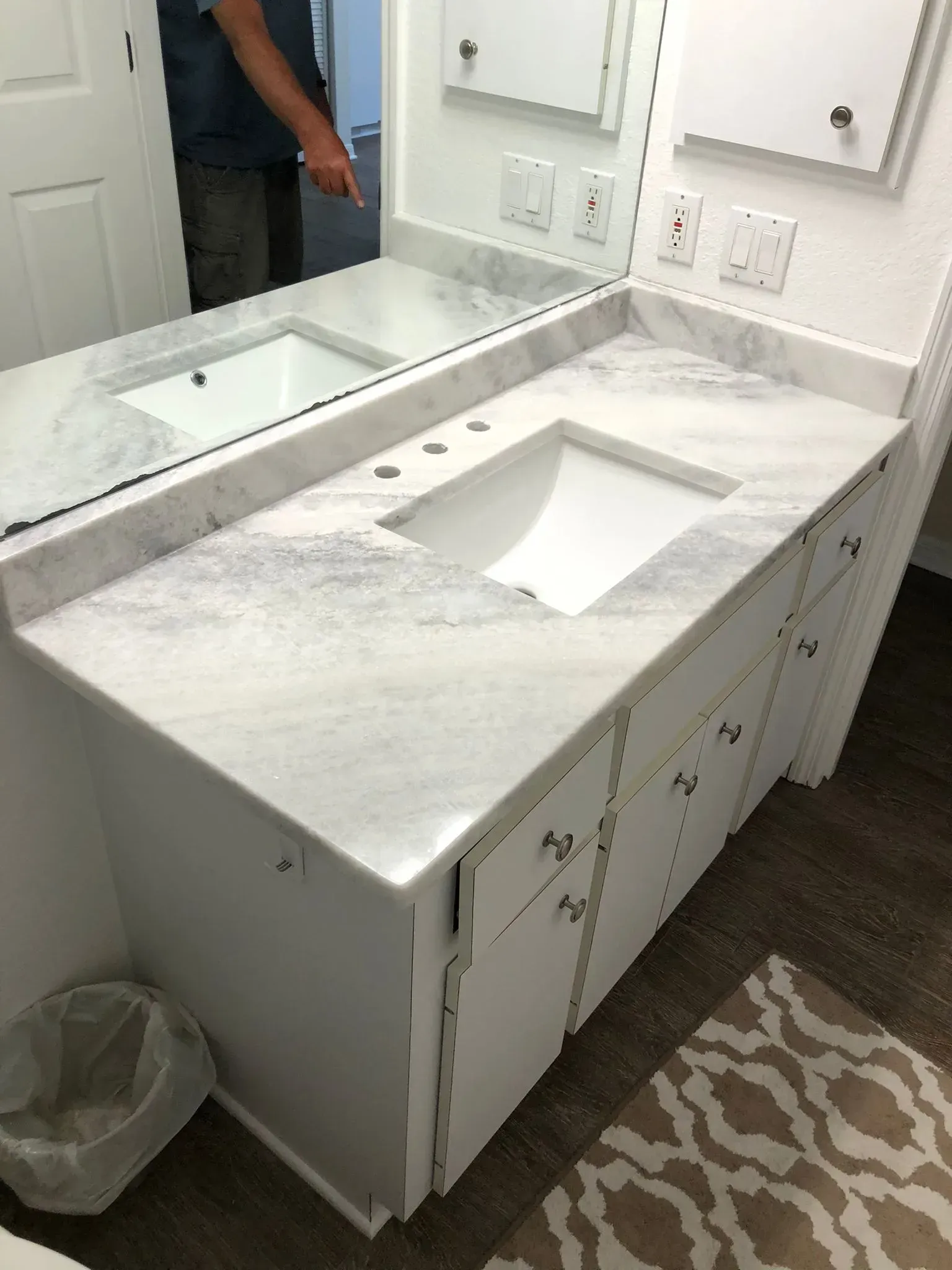 Bathroom vanity with white marble countertop, sink cutout, and a mirror above a dark floor mat.