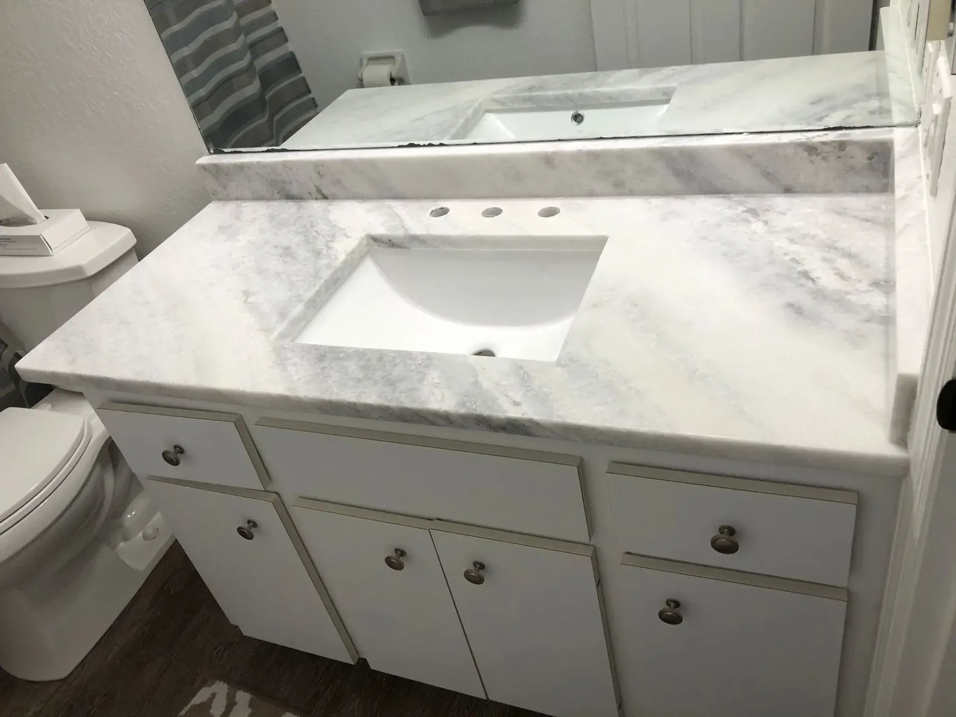 White marble bathroom vanity with sink, mirror, and cabinets, beside a toilet.