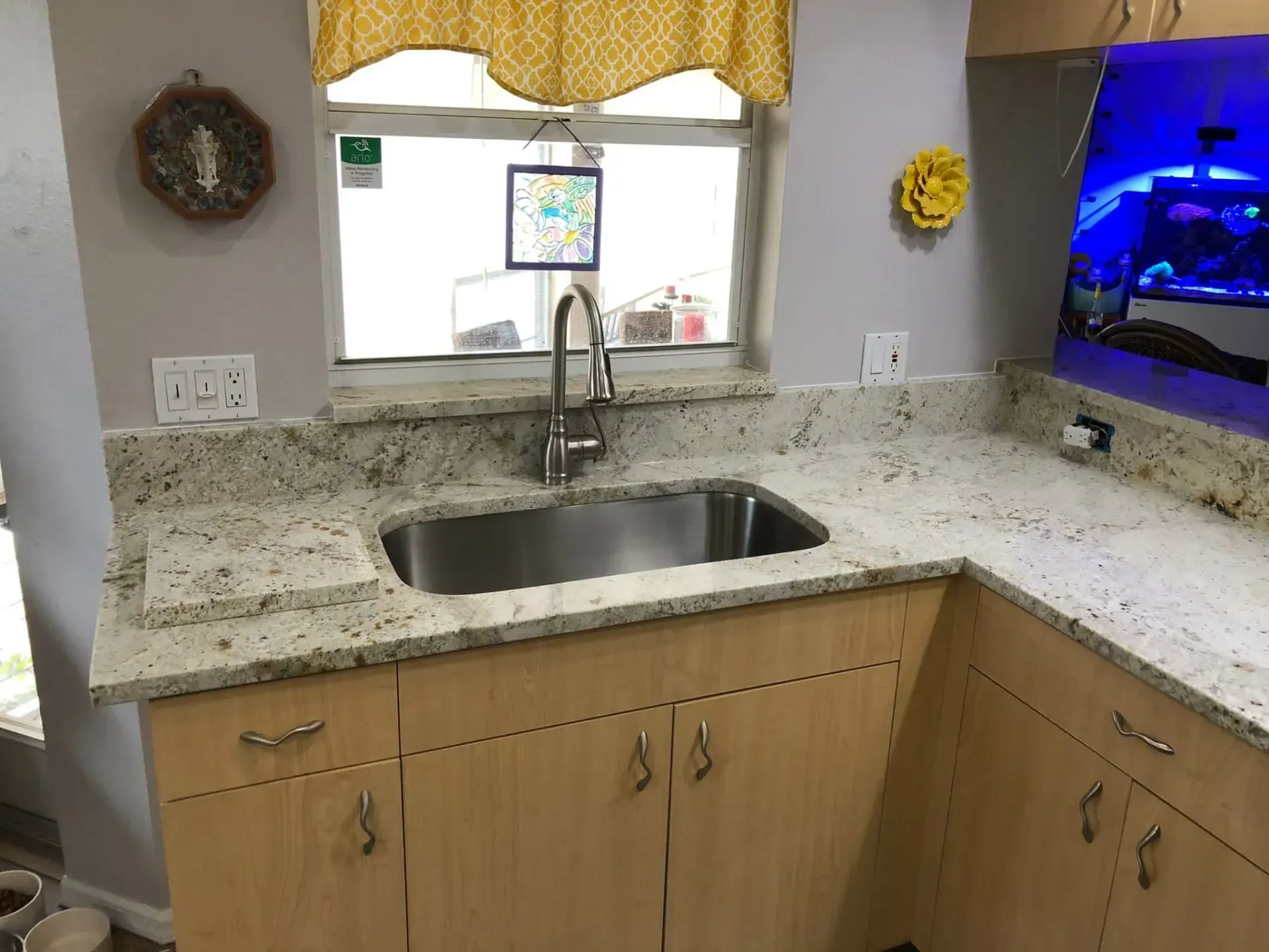 Kitchen sink under a window with granite countertop, light wood cabinets, and blue-lit aquarium on the right.