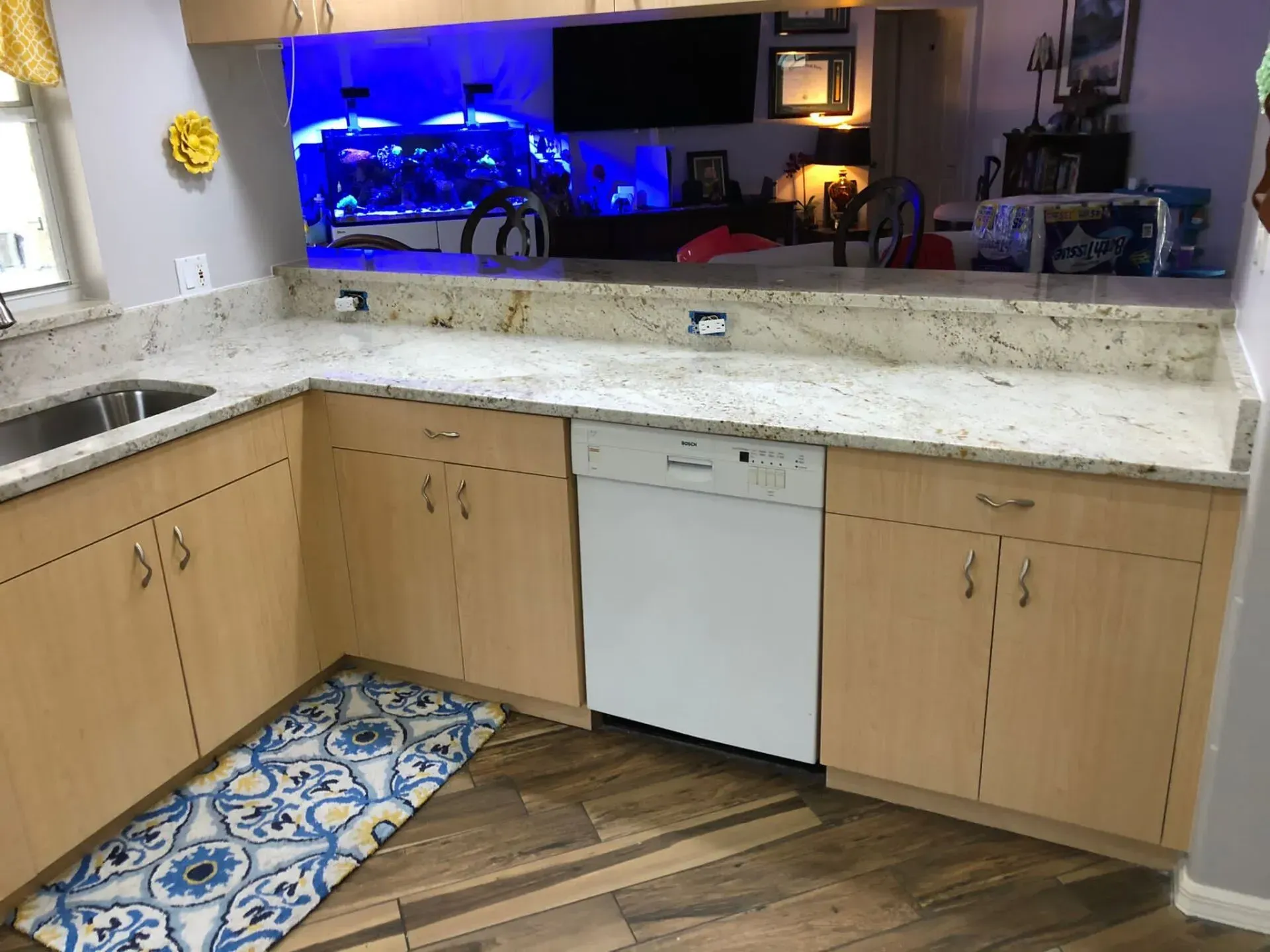 Kitchen with light wood cabinets, white countertop, patterned blue rug, and a view into a blue-lit living room.