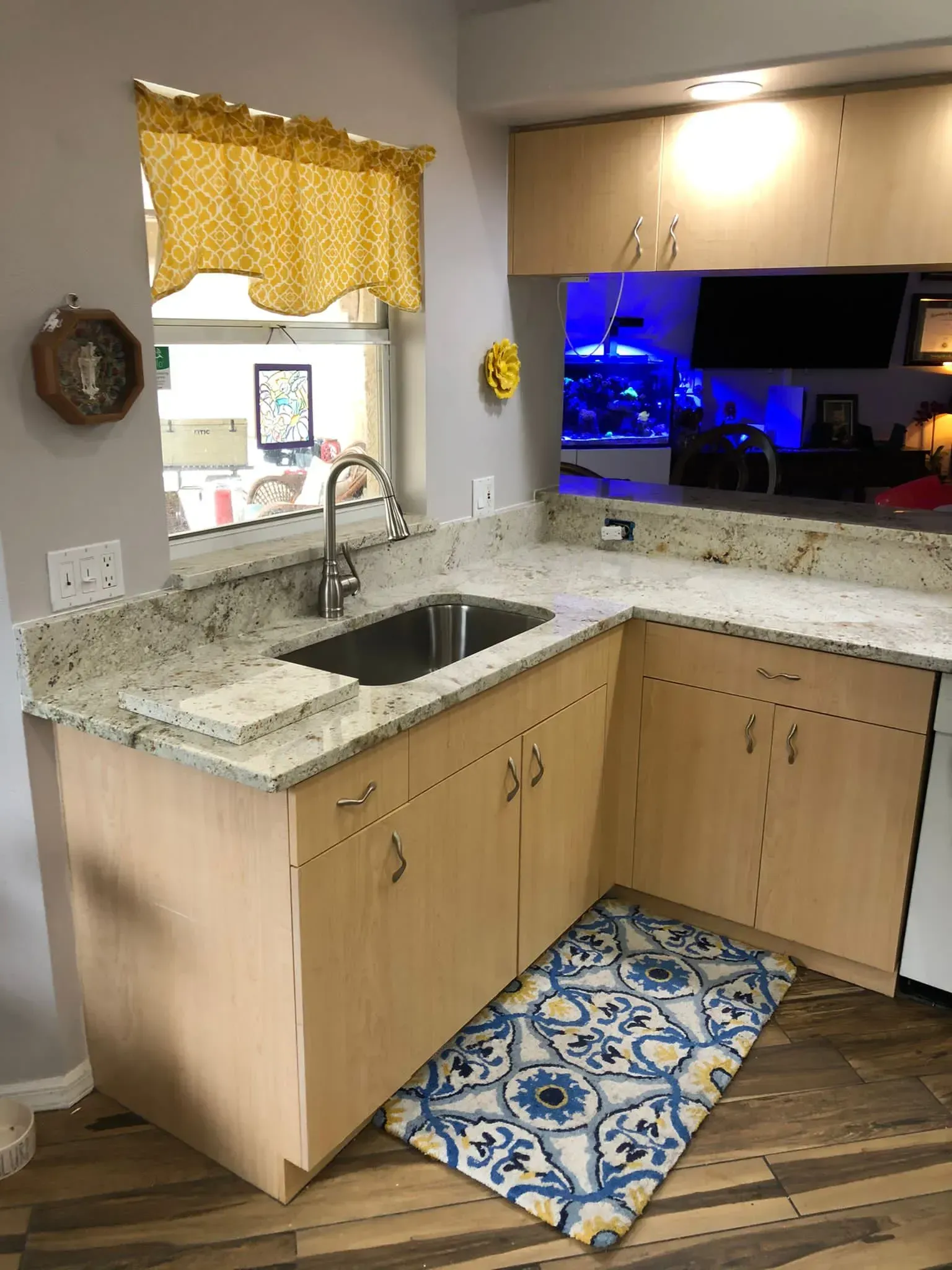 Small kitchen corner with sink, granite counters, wood cabinets, and a blue patterned rug by a window.