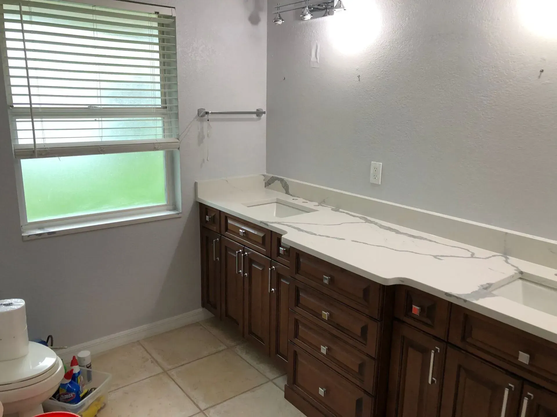 Bathroom with white countertop, dark wood cabinets, beige tile floor, and window with blinds.