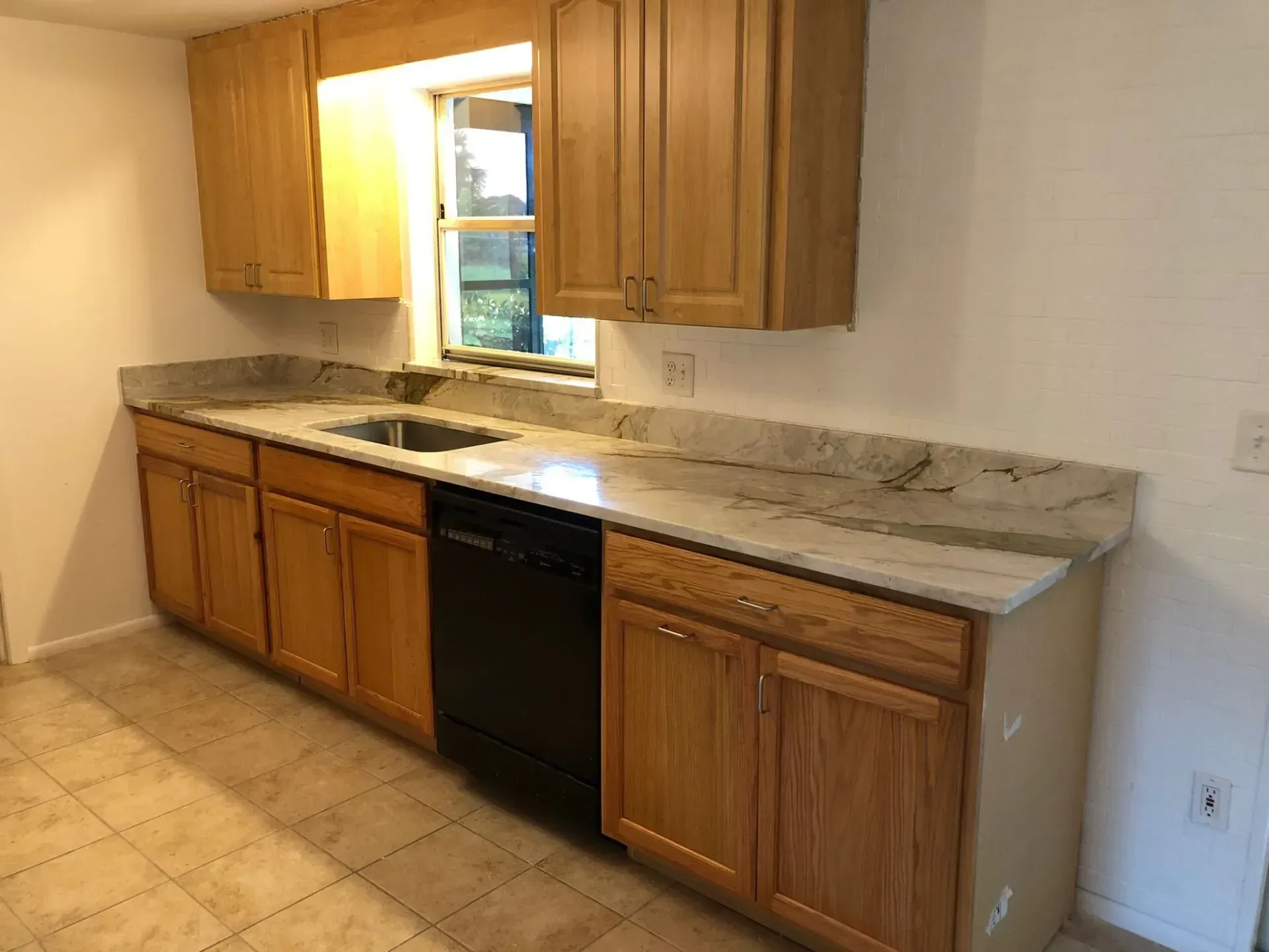 Empty kitchen wall with wood cabinets, granite countertop, black dishwasher, and window above the sink.