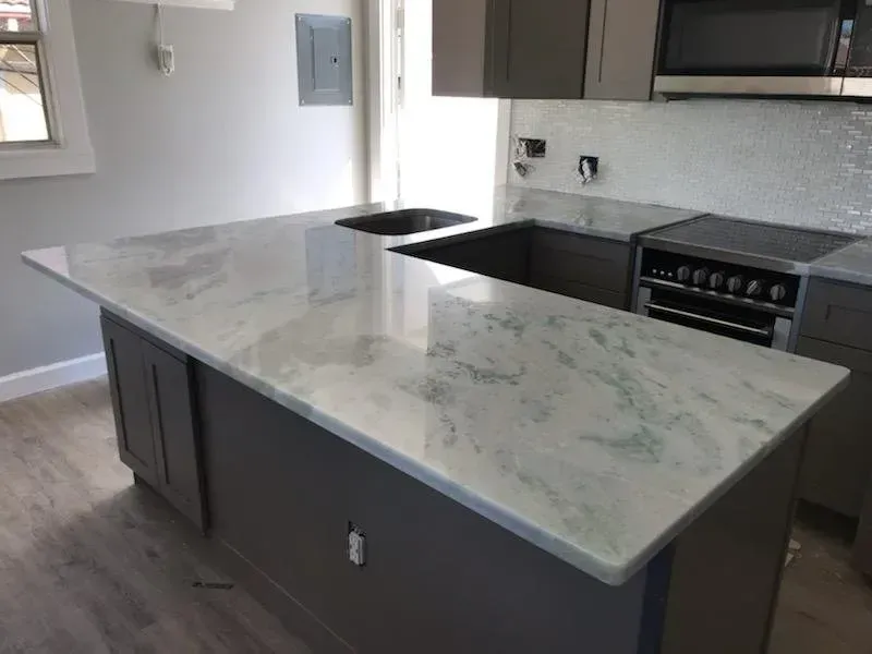 Modern kitchen with large island and white marble countertop, dark cabinets, and built-in oven and sink.