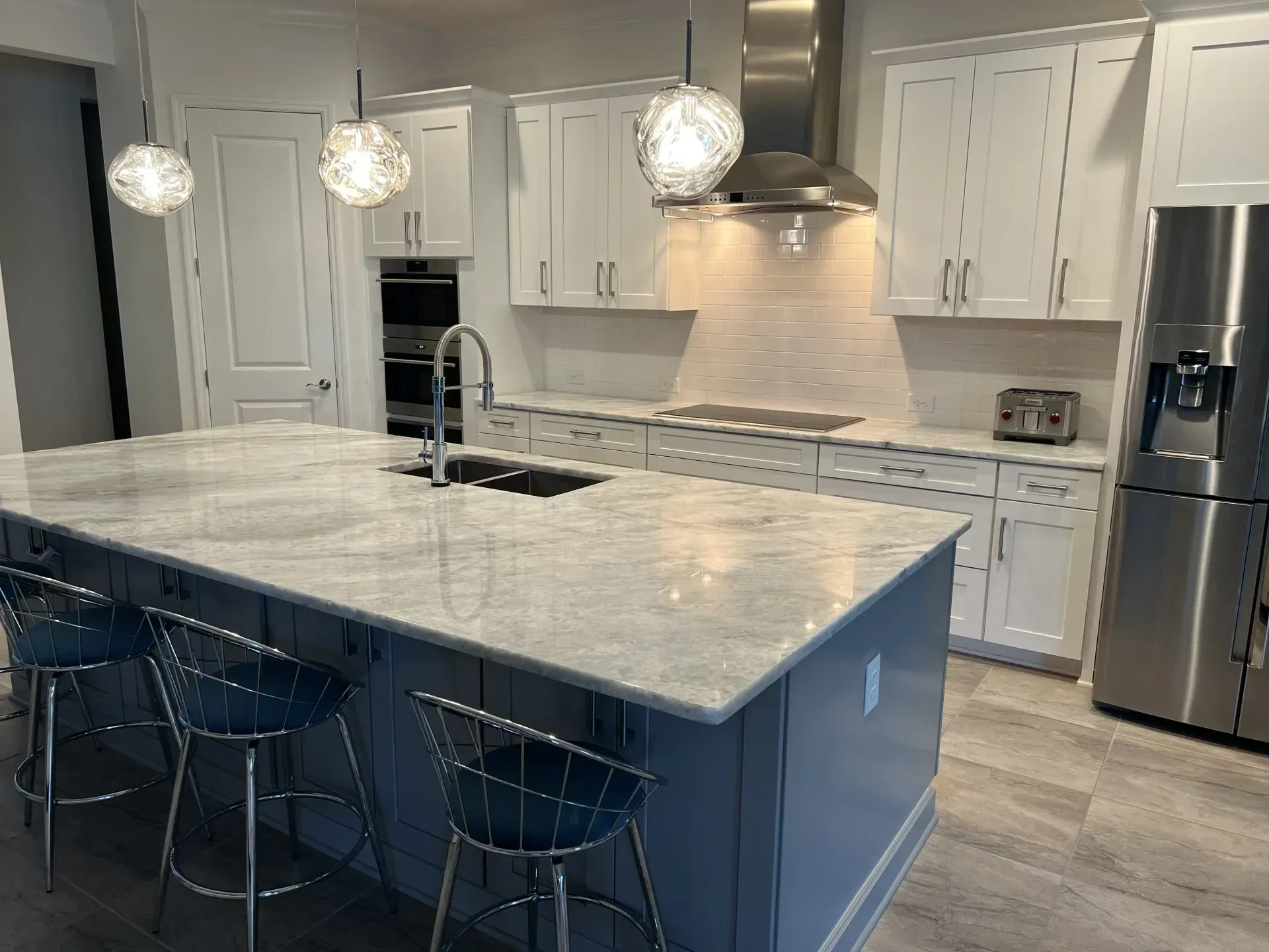 Modern white kitchen with marble island, pendant lights, stainless appliances, and blue barstools