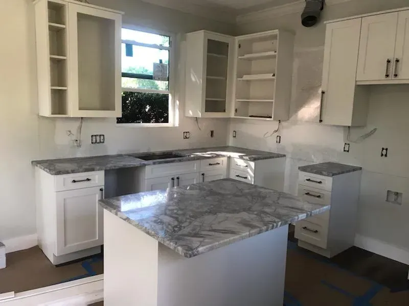 Bright kitchen with white cabinets, marble countertops, and a central island under a window.