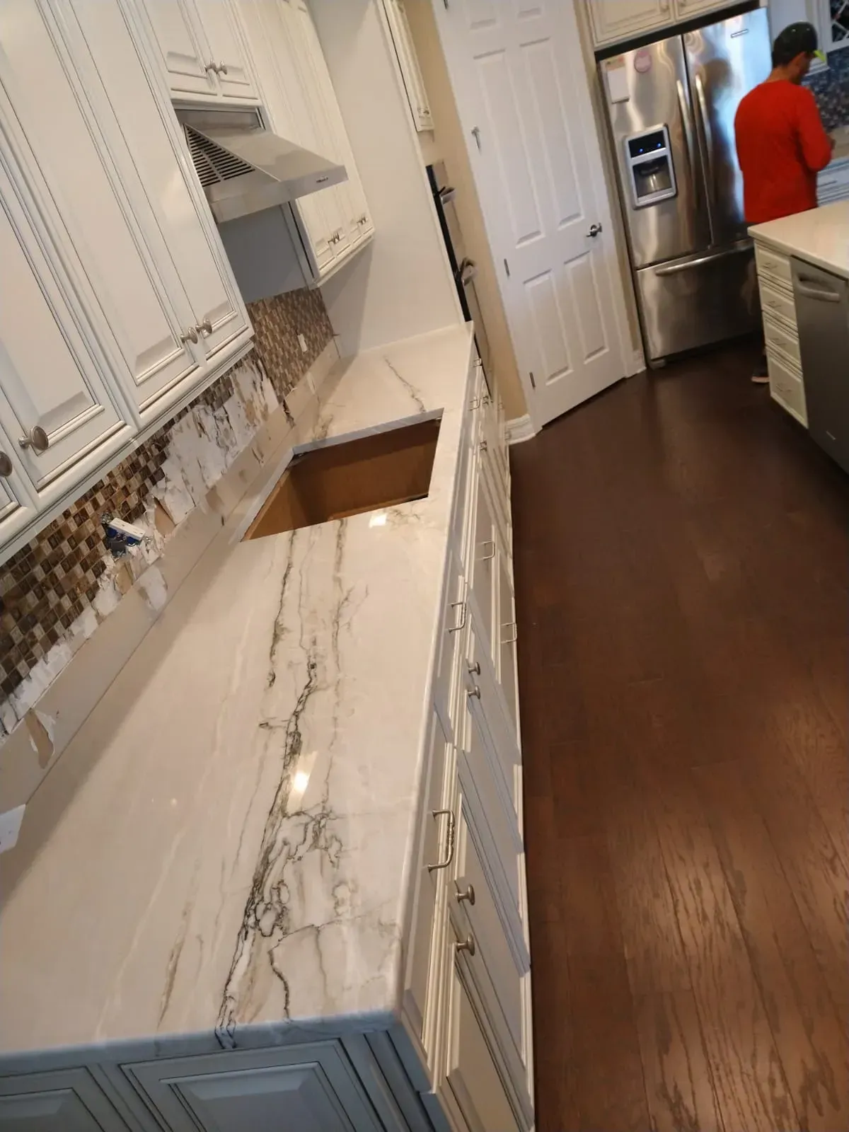 Kitchen with white cabinets and marble countertop; a person in a red shirt stands near the hallway.