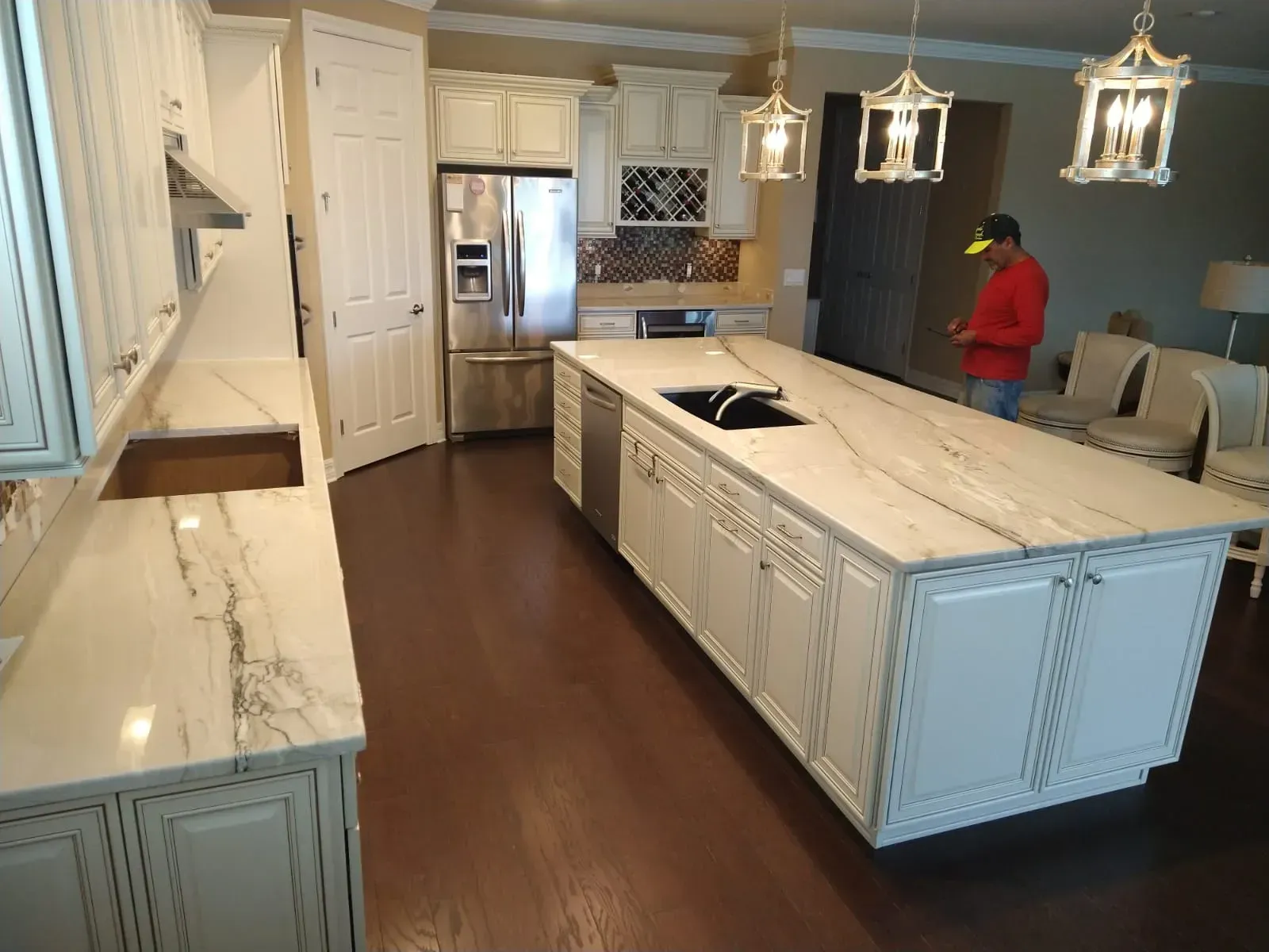 Bright kitchen with large island, white cabinets, dark wood floors, and a person in a red shirt near the back.