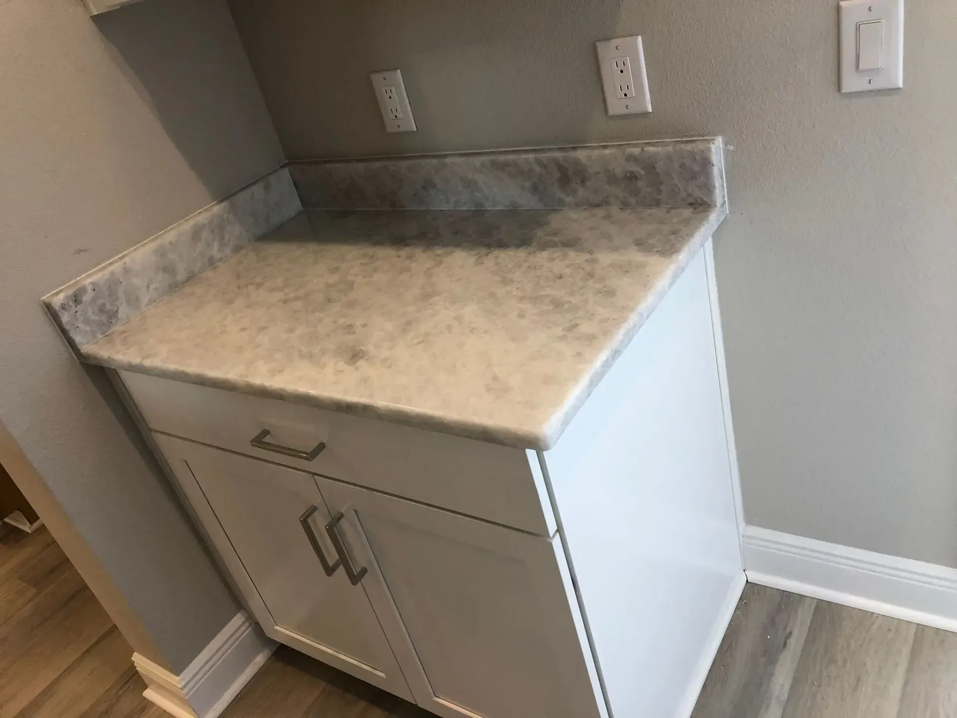 Small white cabinet with marble-look top in a corner under wall outlets, likely a laundry or utility area