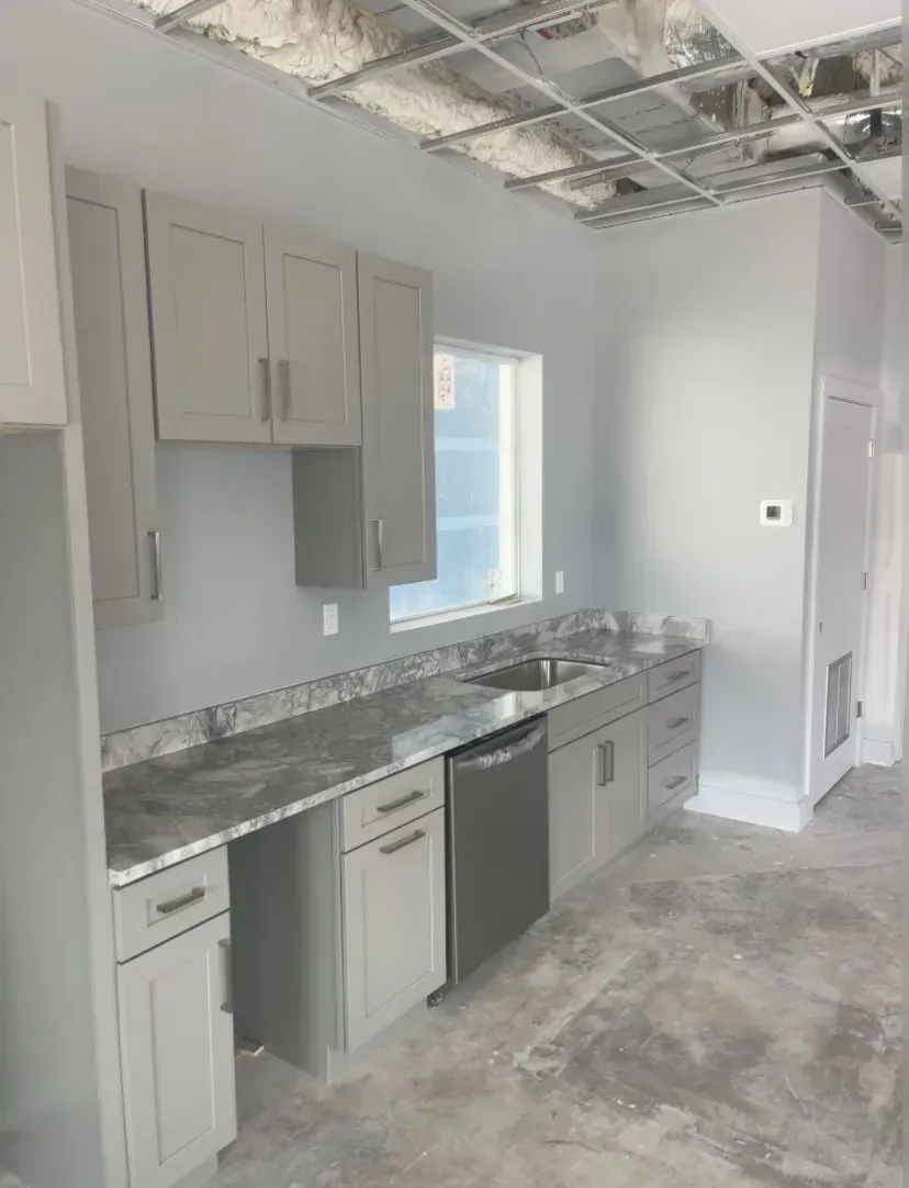 Unfinished kitchen with gray cabinets, marble-look countertops, stainless dishwasher, and exposed ceiling.