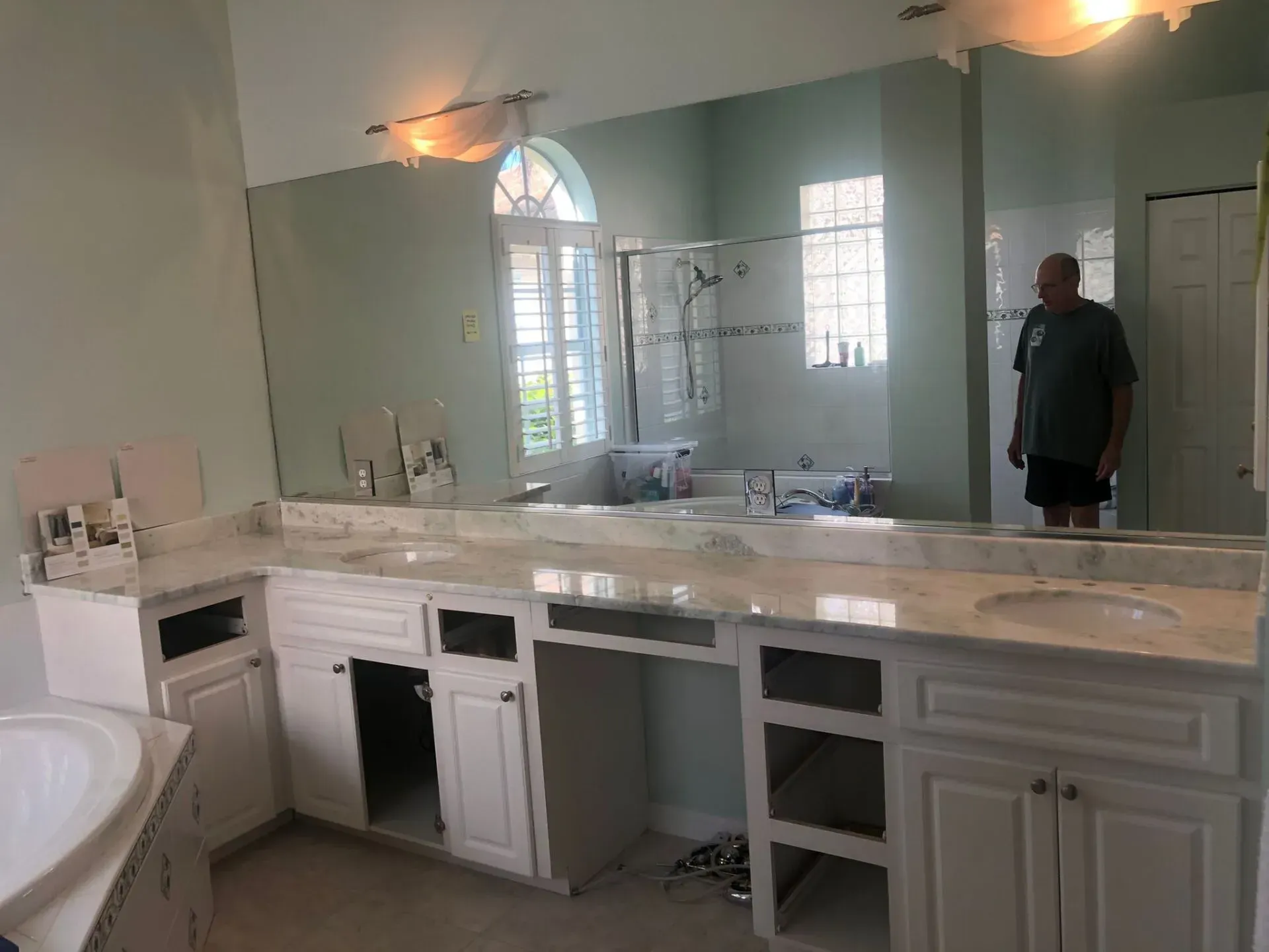 Bathroom with white vanity under renovation, mirrors and lights, and a man standing by the wall.