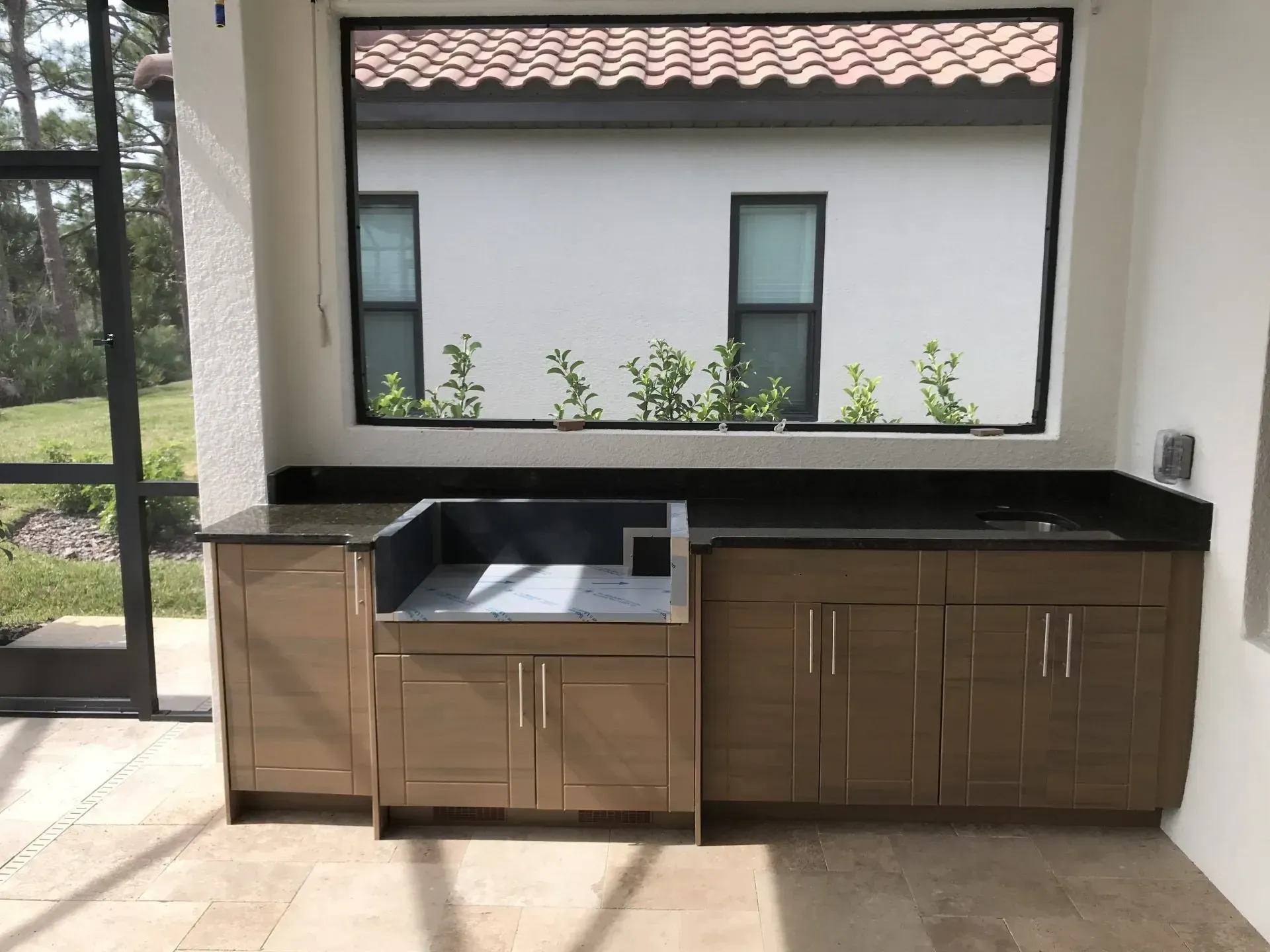 Outdoor built-in kitchen with brown cabinets, black countertop, and sink under a windowed opening
