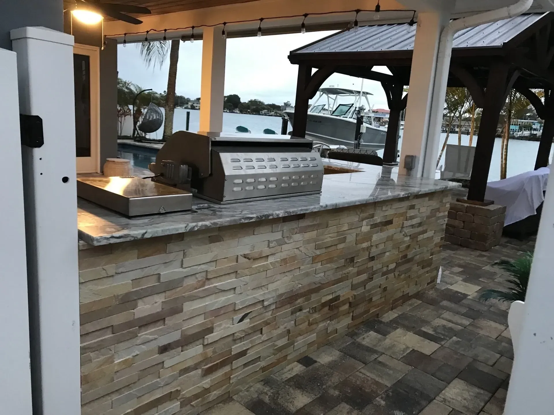 Outdoor bar with stone counter, grill, and waterfront gazebo seating at sunset