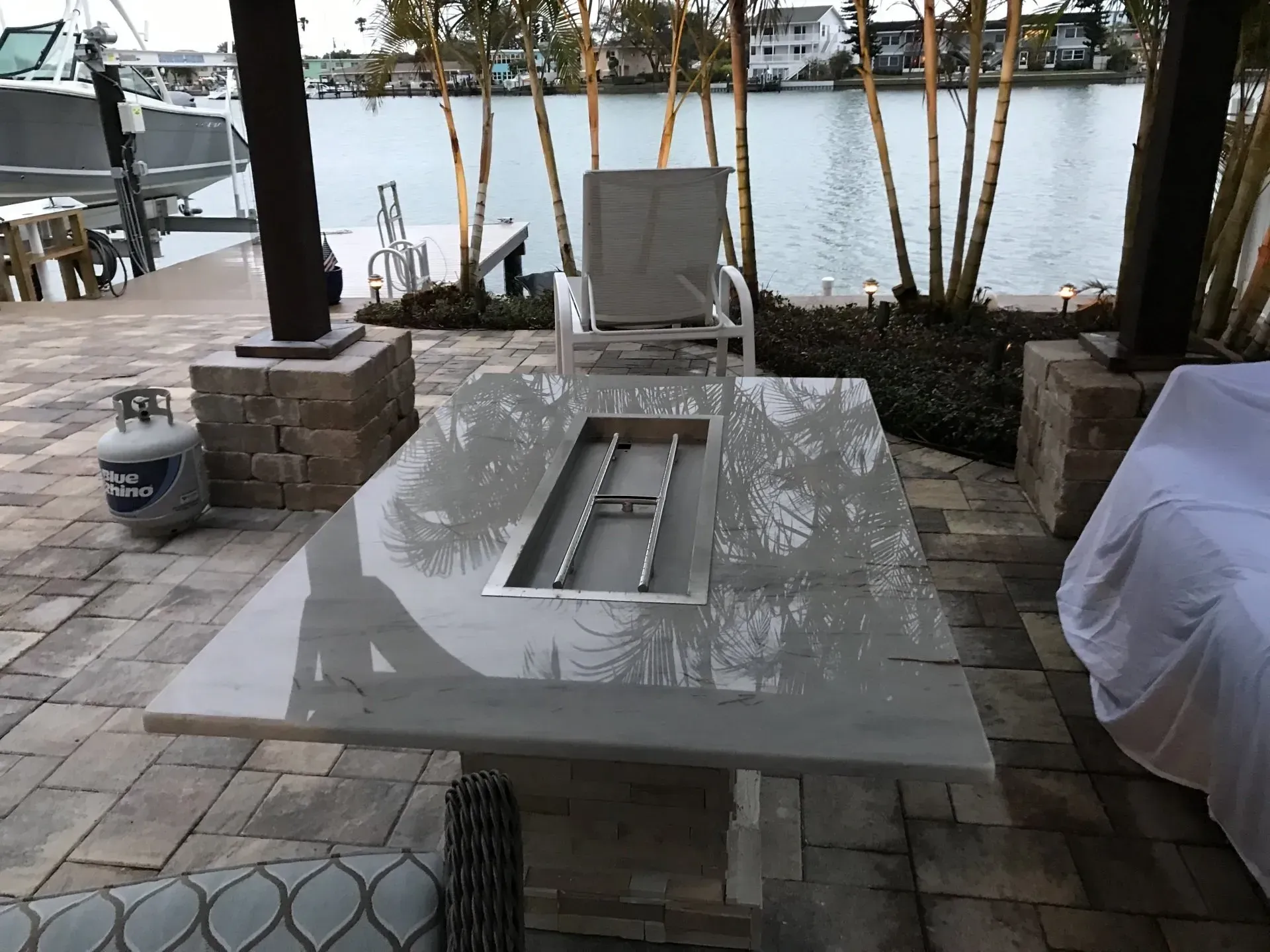 Outdoor stone fire pit table beside a lake with chairs and covered furniture on a patio