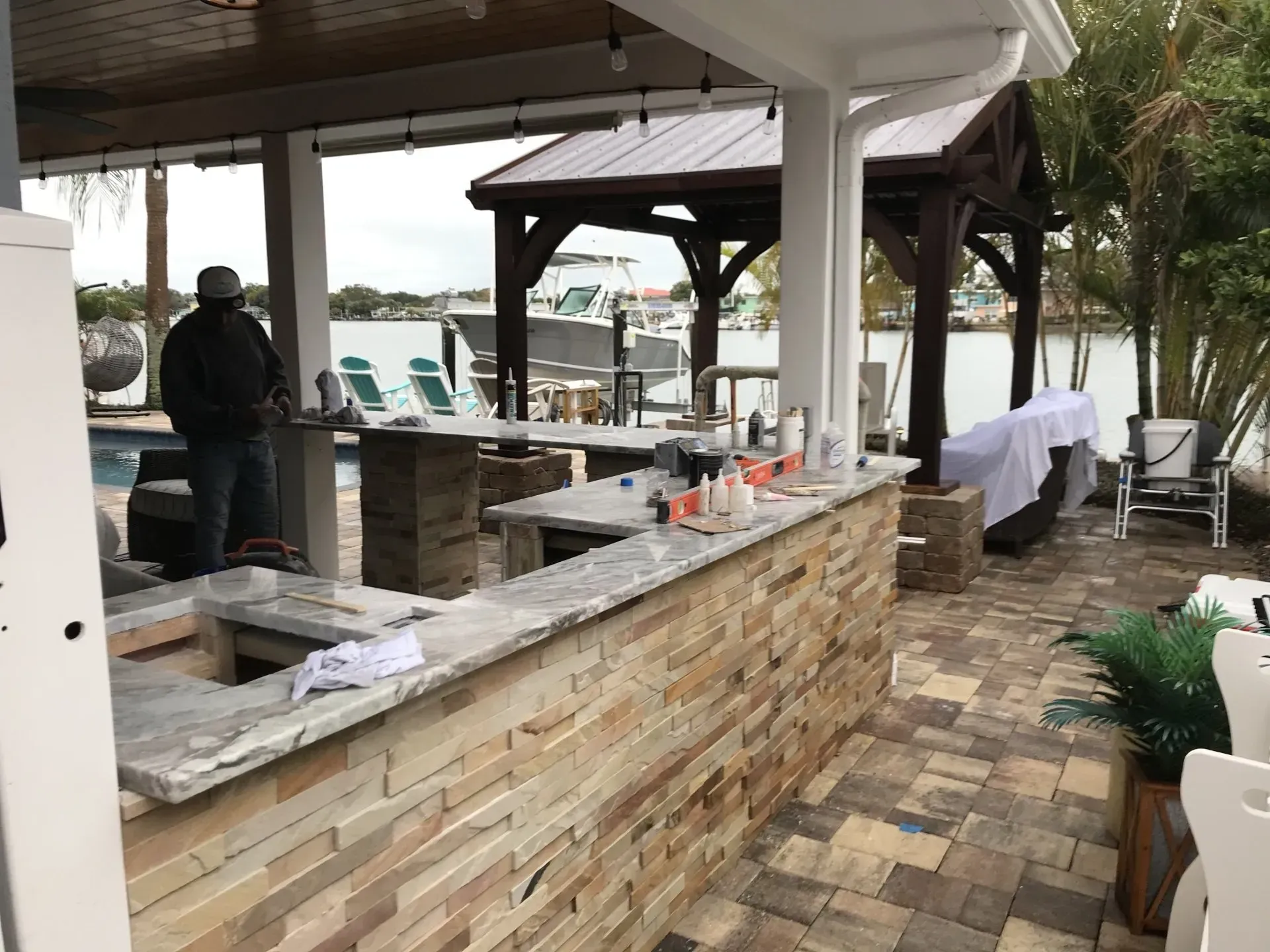 Covered outdoor patio with stone bar, gazebo, chairs, and a person standing near the grill.